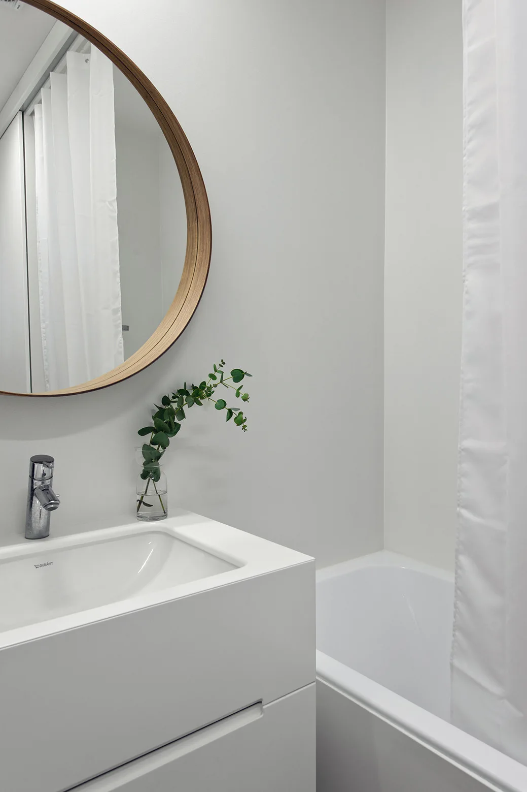 A minimalist white bathroom vanity with an integrated sink, a chrome faucet, a round wooden-framed mirror, and a white bathtub with a curtain.