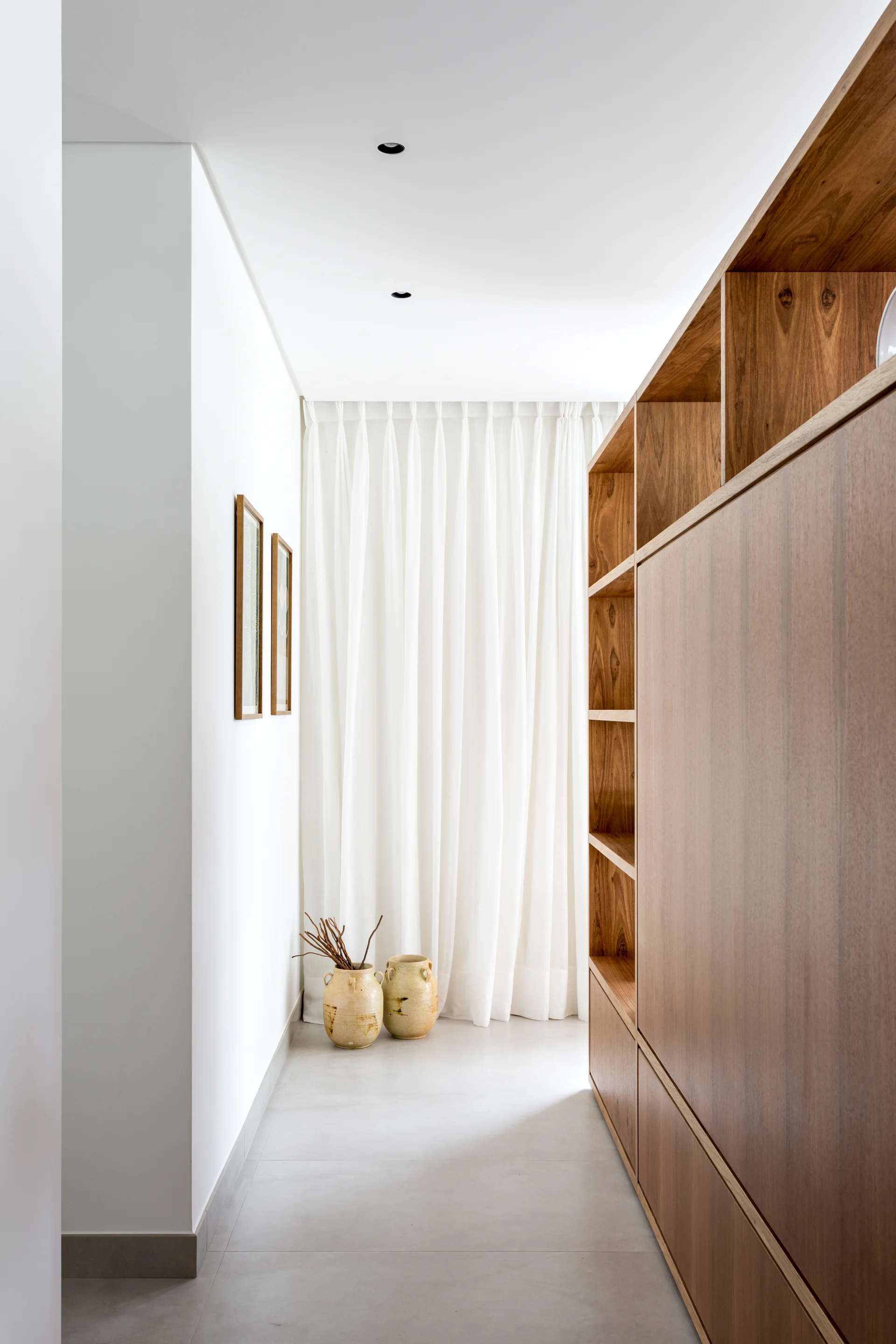 A minimalist hallway features custom wood cabinetry along one wall, two framed pictures, and white floor-to-ceiling curtains at the far end.