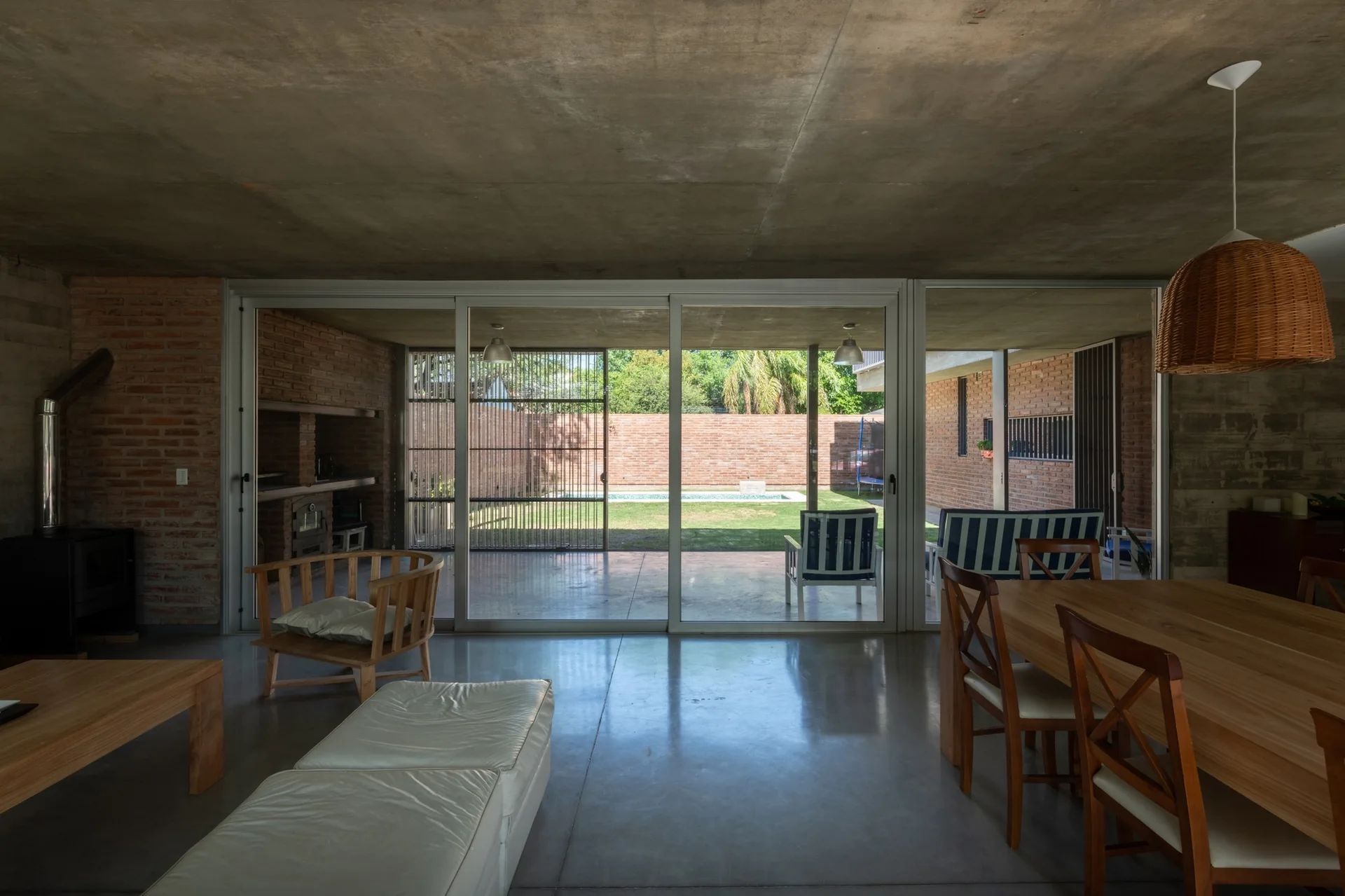 An open-plan living and dining room features an exposed concrete ceiling, brick walls, a wooden dining table, and large sliding glass doors opening to a yard.