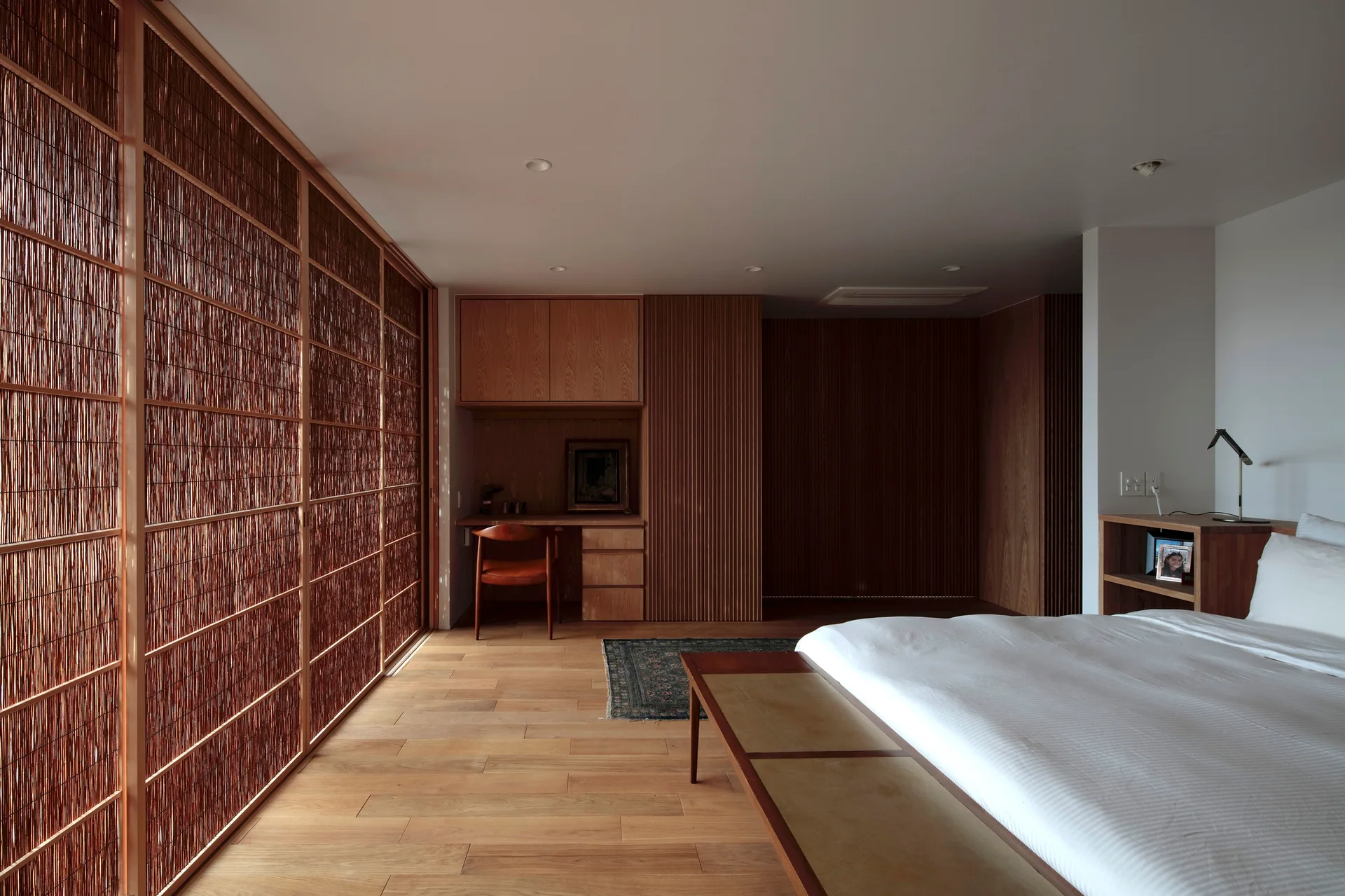 A minimalist bedroom featuring warm wood floors, a large reed sliding screen, an integrated wooden desk nook, and a bed with white linens.