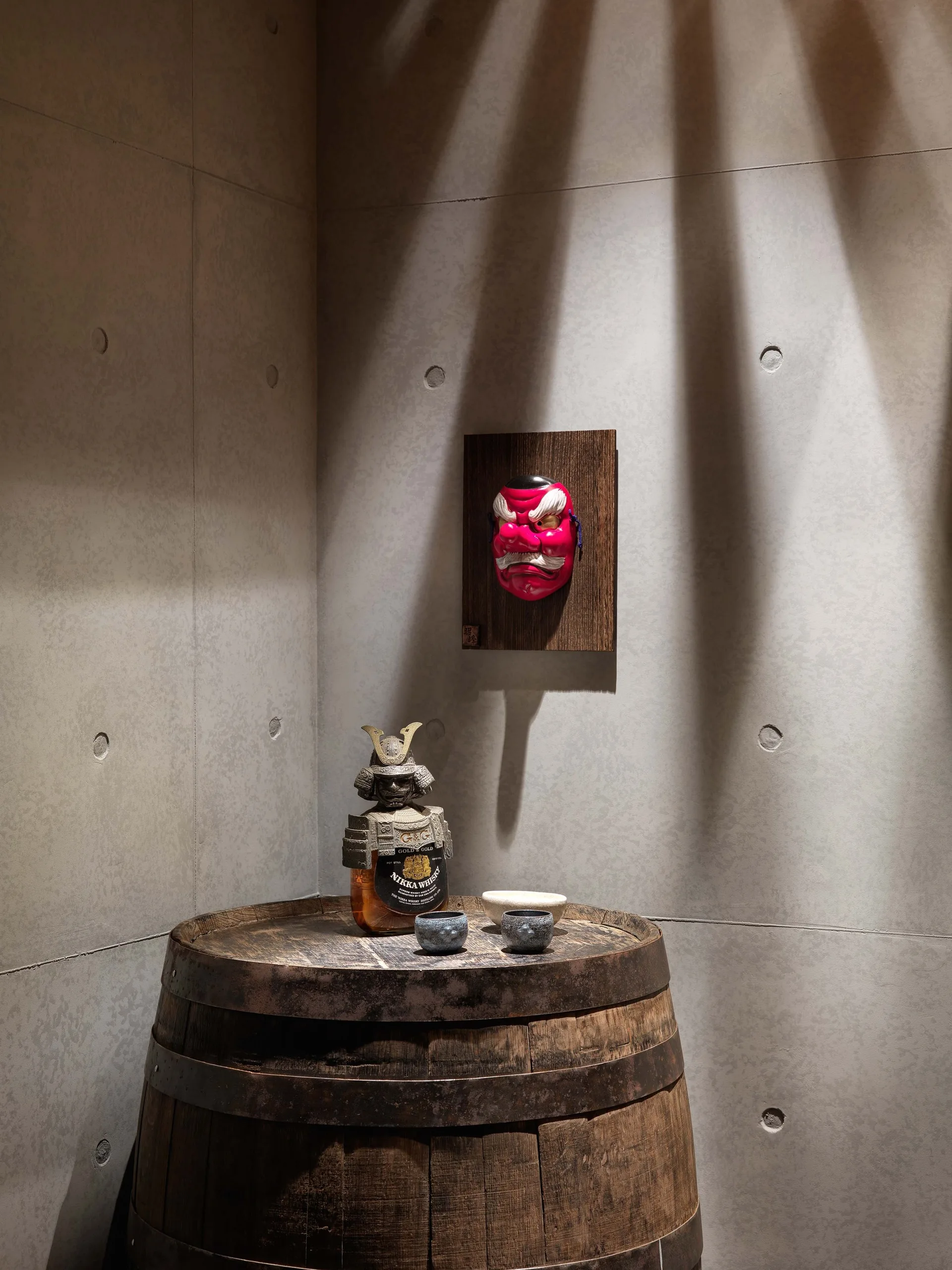 A decorative corner vignette features a wooden barrel used as a table beneath a red Japanese mask on a grey concrete wall.