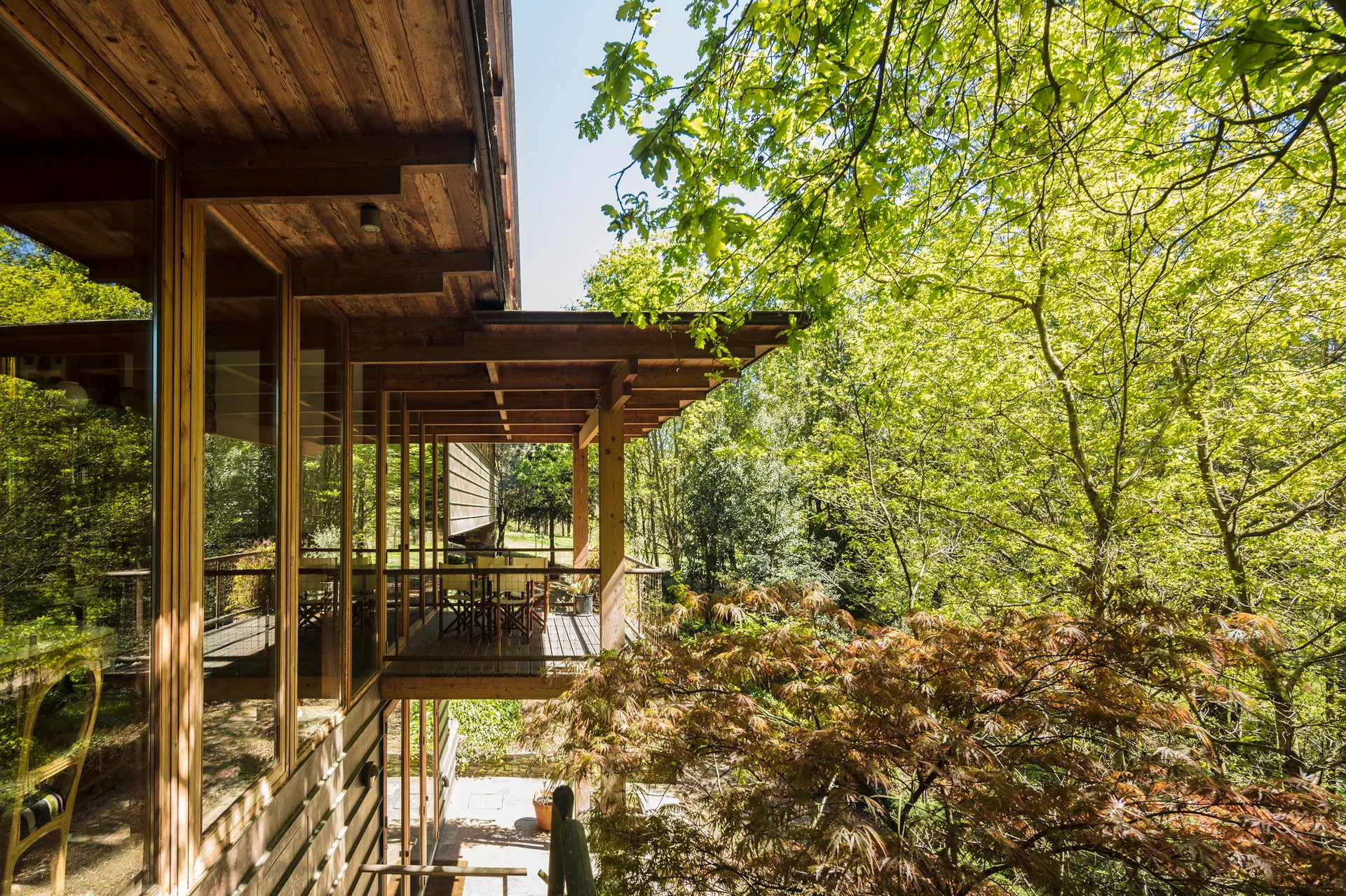 An elevated wooden deck features an outdoor dining table, glass doors, and a deep roof overhang overlooking a dense forest.