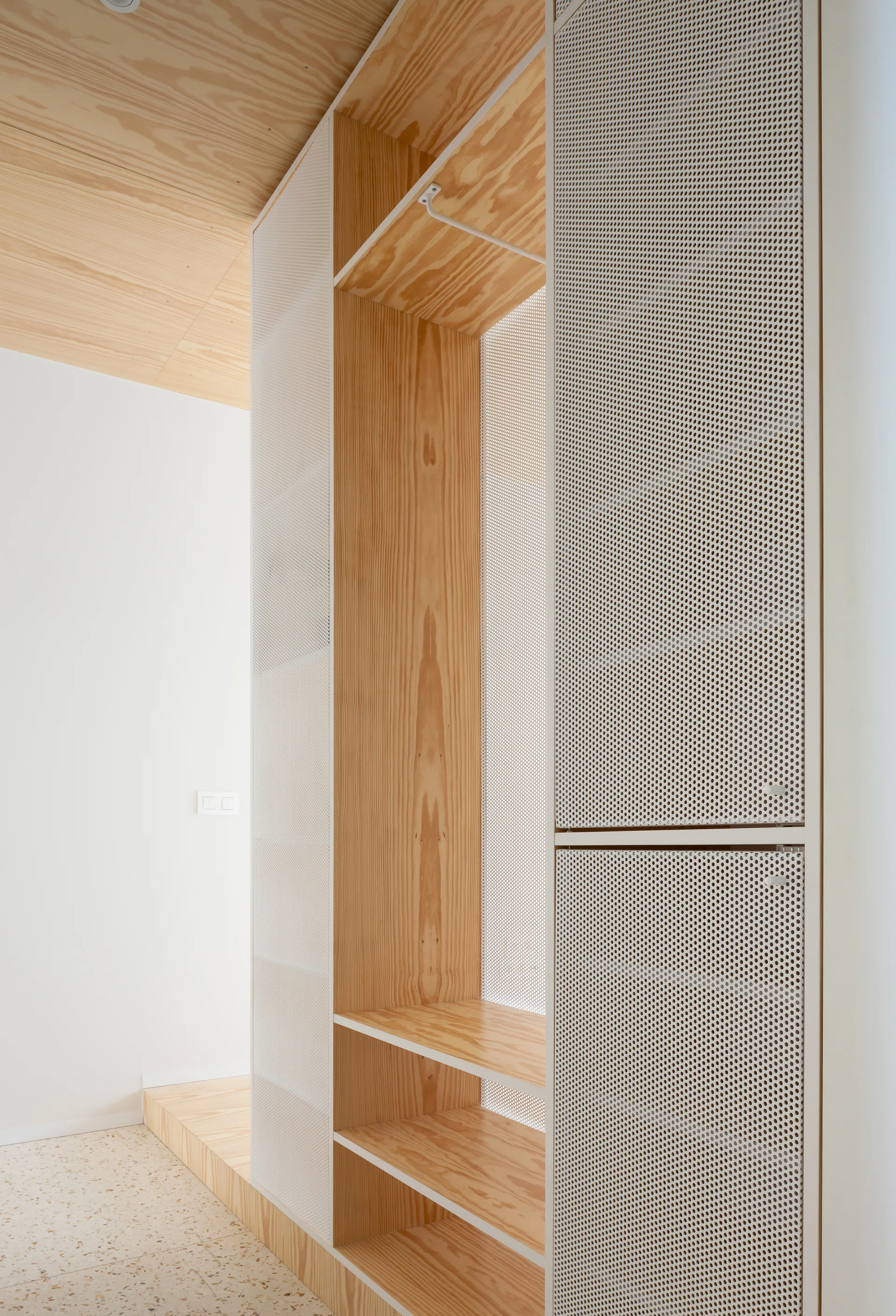 A tall custom plywood storage unit with white perforated metal doors sits beneath a matching plywood ceiling on a speckled terrazzo floor.