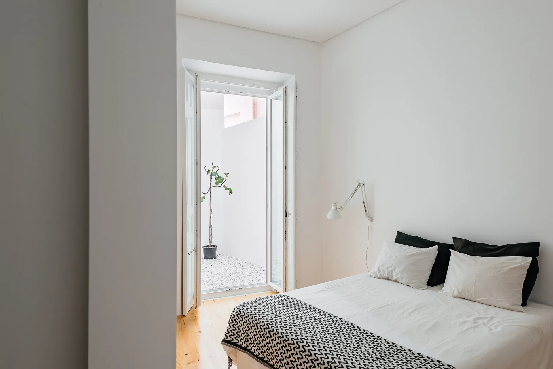 A minimalist white bedroom features a bed with black and white linens next to glass double doors that open to a small gravel courtyard with a plant.