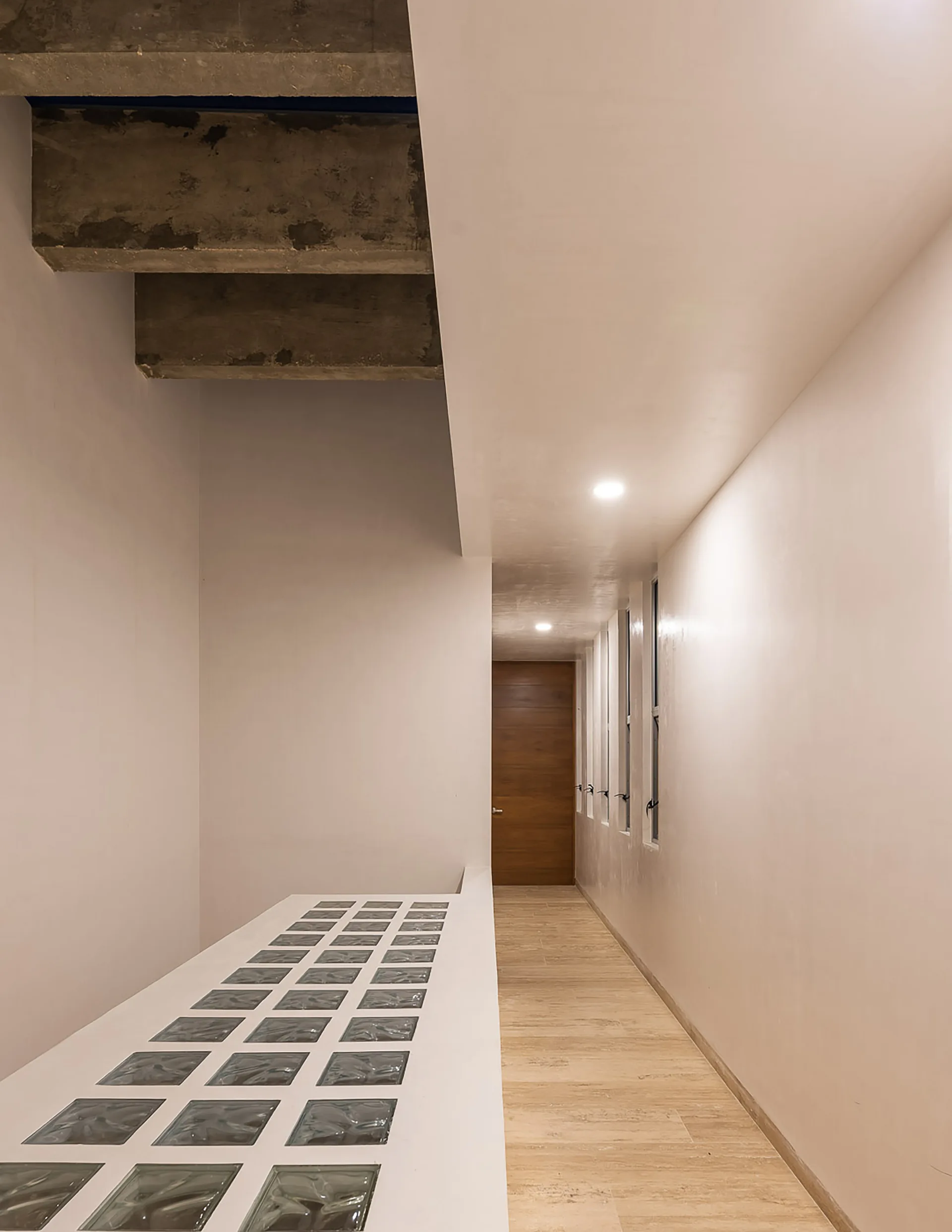A long minimalist hallway features white walls, a tiled floor, a glass block floor insert, and exposed concrete beams on the ceiling.