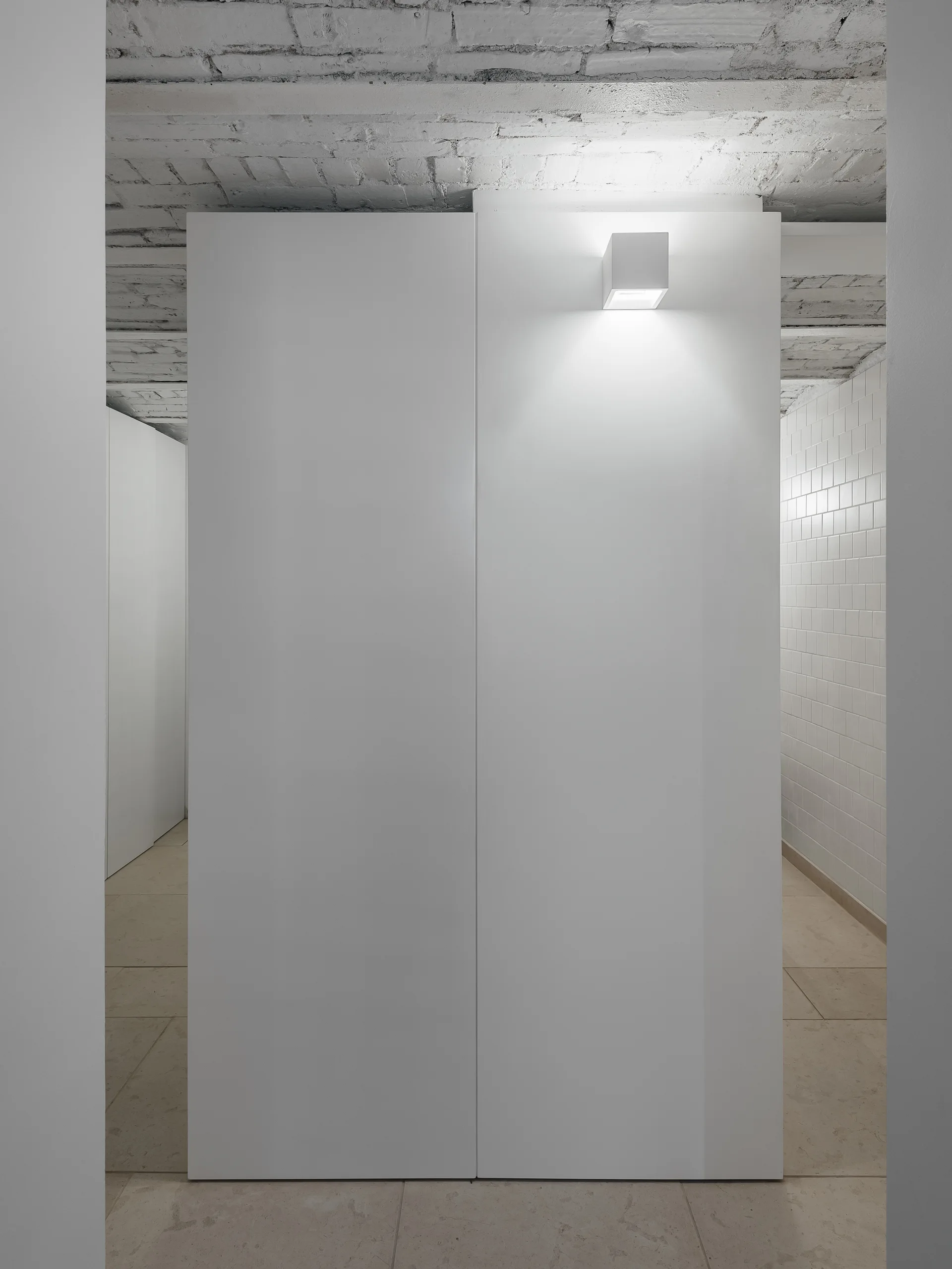 A white floor-to-ceiling partition wall stands in a minimalist hallway with an exposed painted brick ceiling and a small square light fixture.