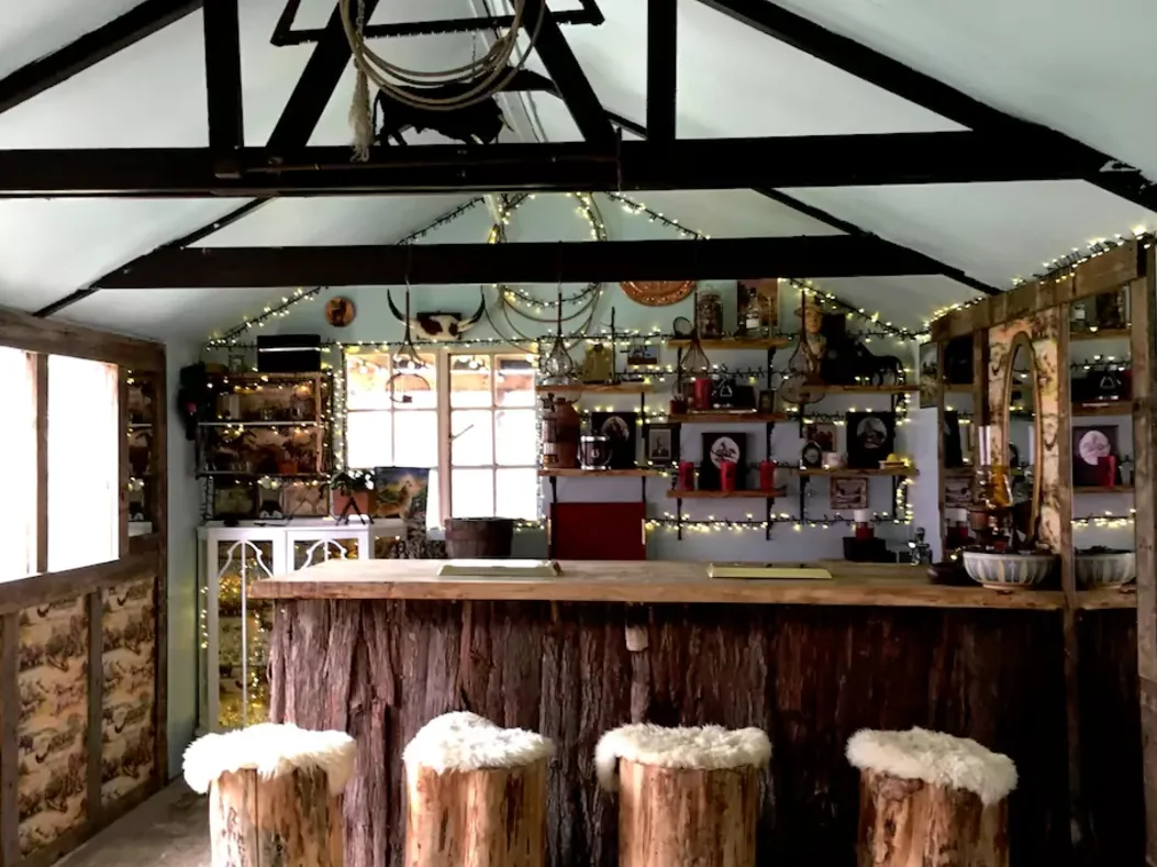 A rustic home bar featuring a tree-bark counter base, log stools topped with white fur, and wall shelves decorated with warm string lights.