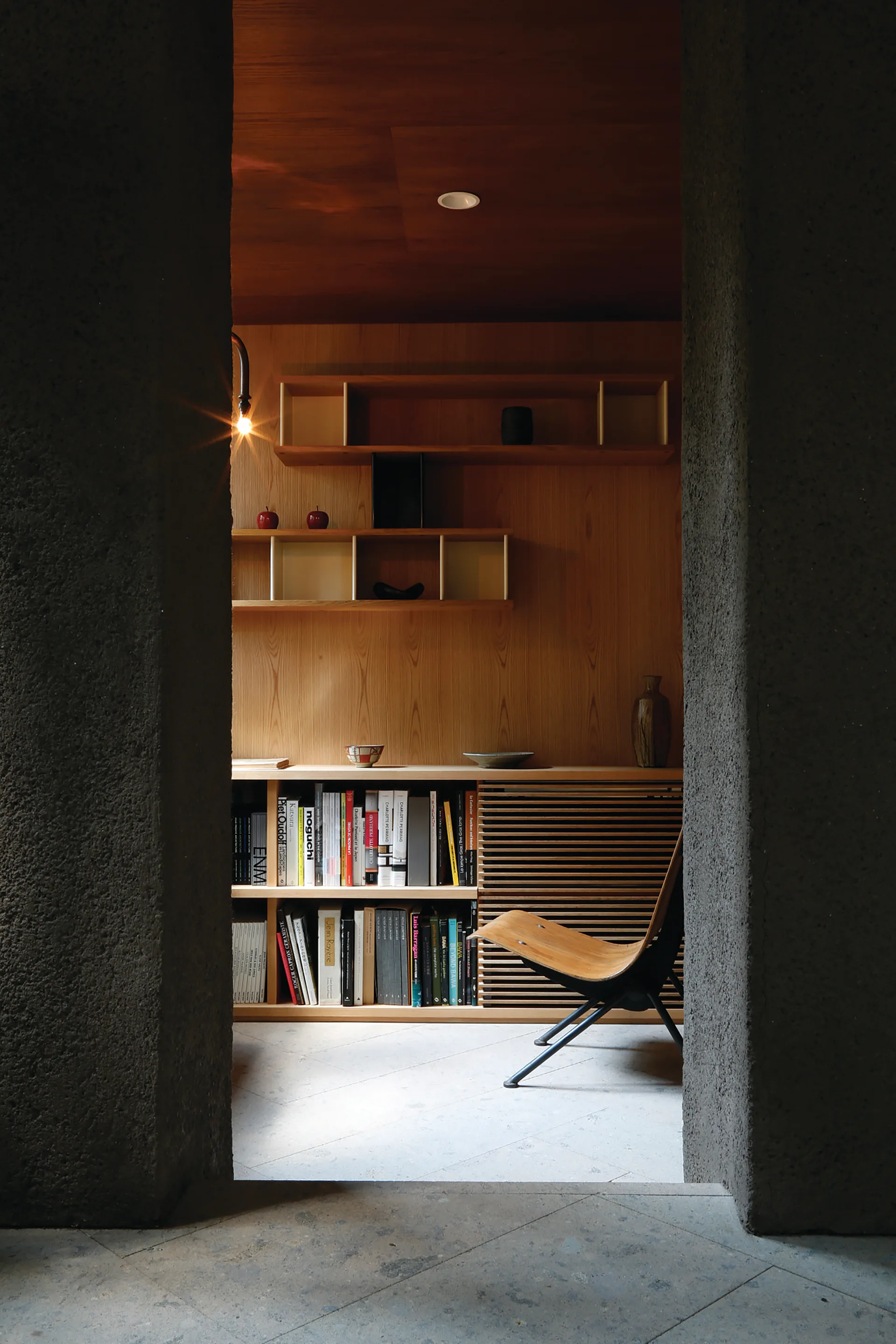 A view through a rough dark stone doorway reveals a warm wood-paneled room with built-in bookshelves, a slatted cabinet door, and a wooden lounge chair.