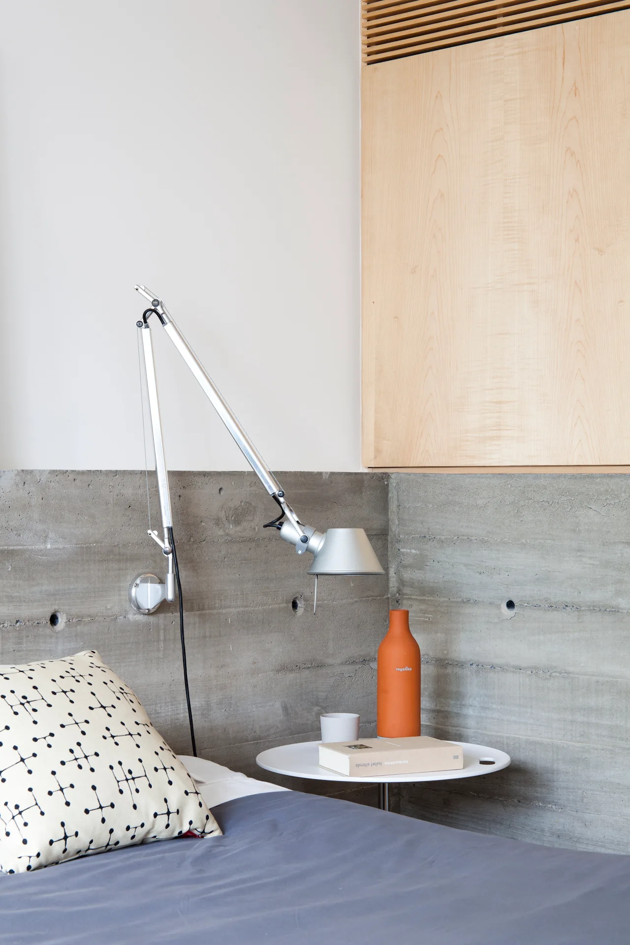 A modern bedside setup features a silver swing-arm lamp mounted to an exposed concrete wall next to a round white table with an orange vase.