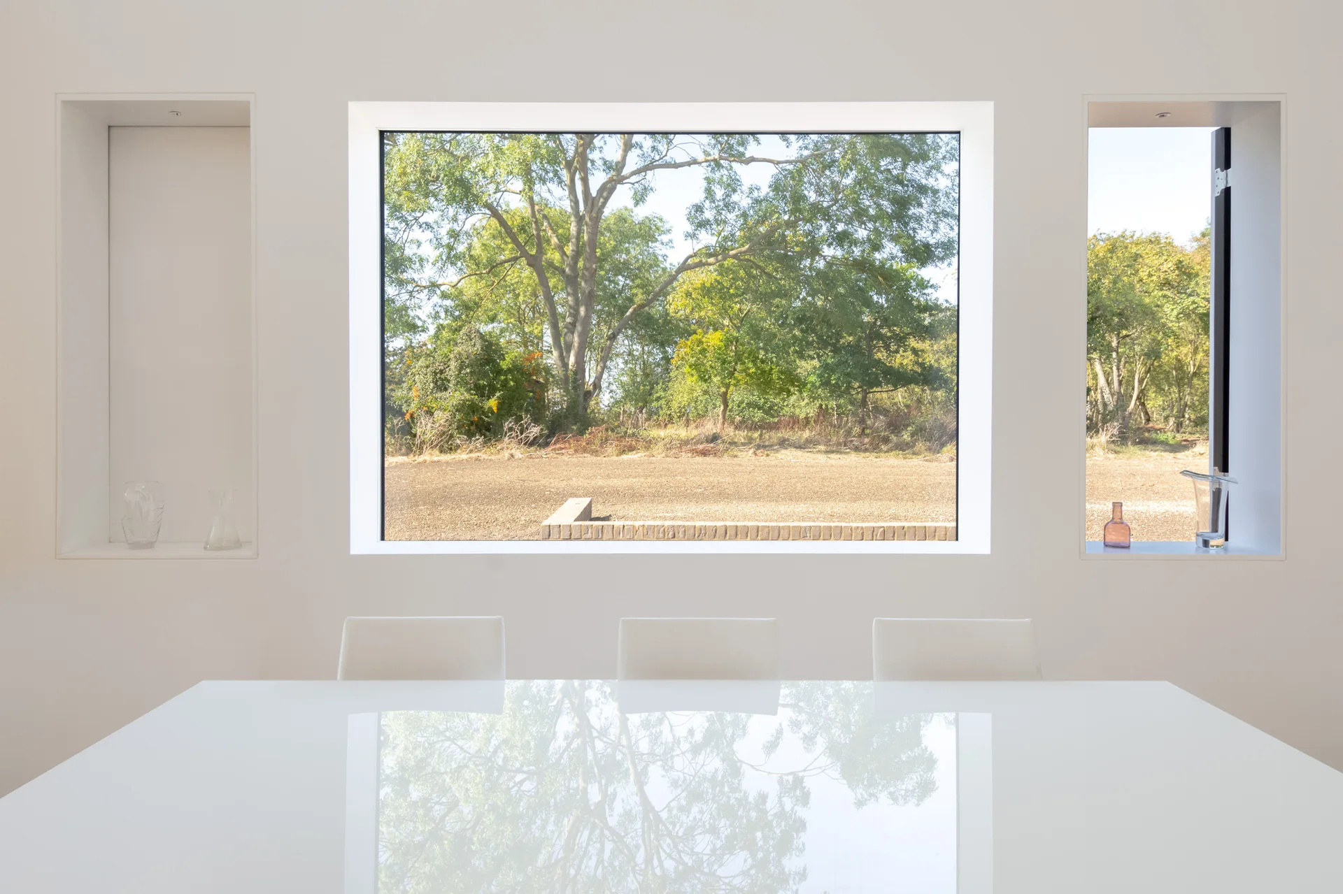 A minimalist white dining room features a high-gloss white table reflecting a large window that frames green trees.