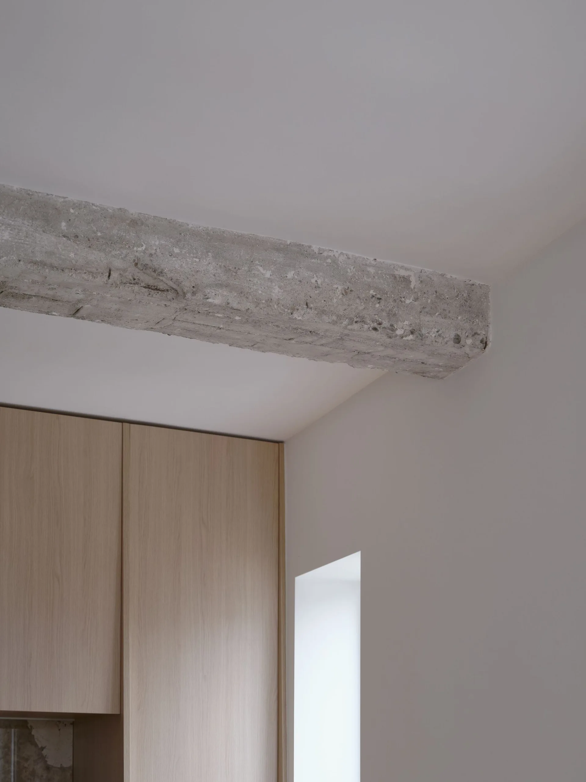 A minimalist interior corner featuring an exposed concrete ceiling beam above flat-front wood cabinets and a recessed white wall niche.