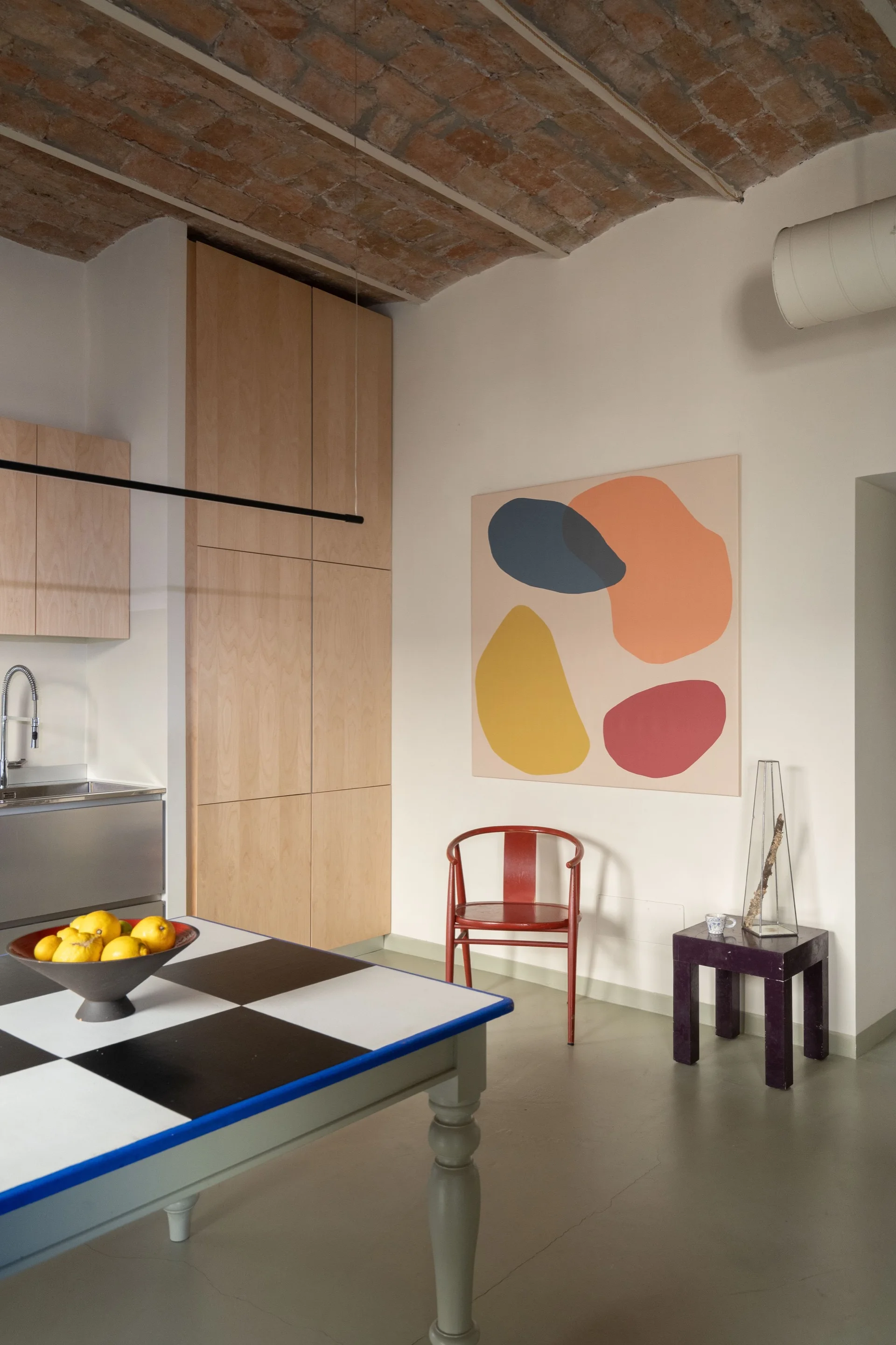 An eclectic kitchen features a checkered dining table, a red chair, an abstract painting, light wood cabinets, and an exposed brick arched ceiling.