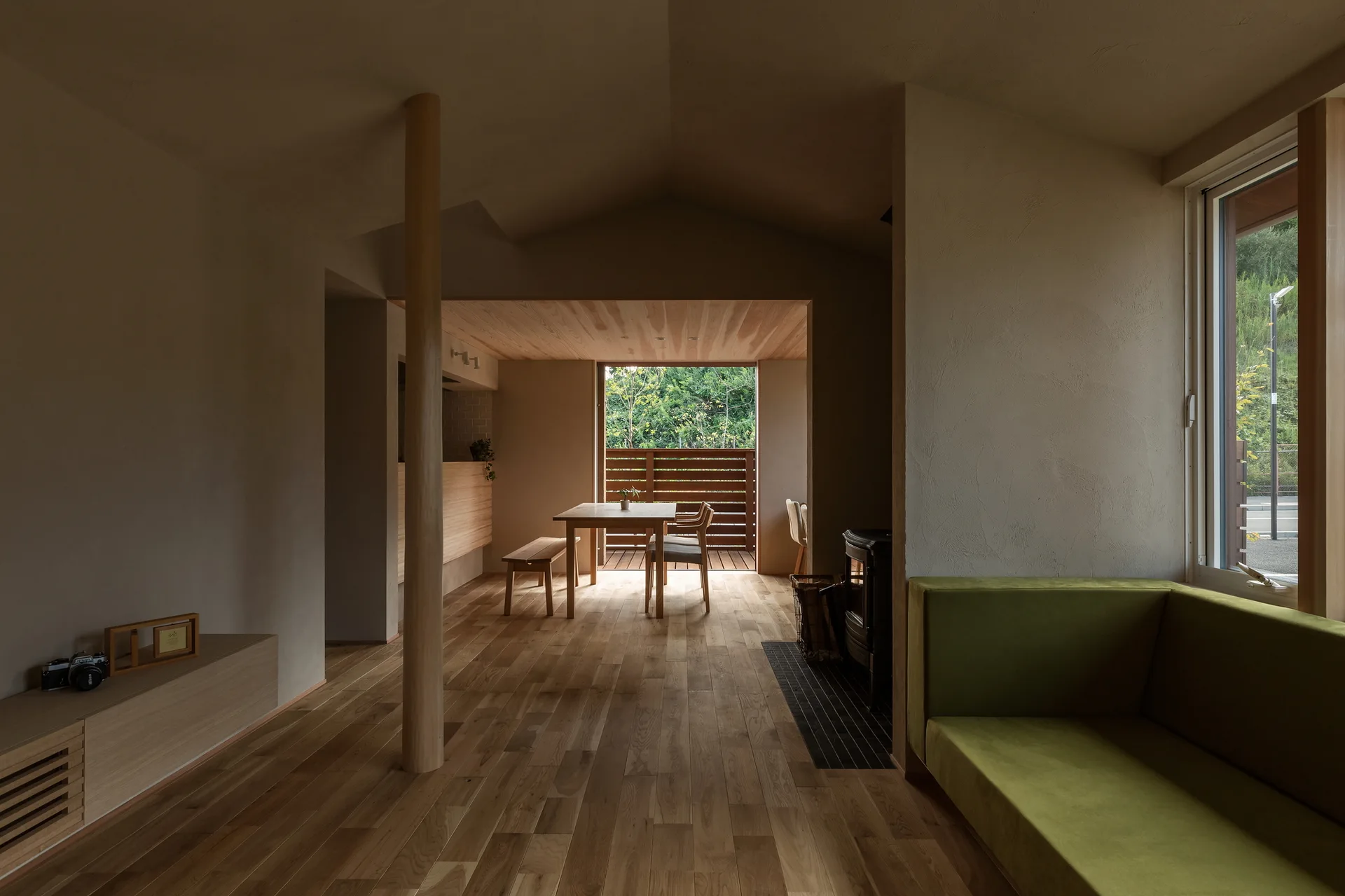 A minimalist living and dining space featuring textured plaster walls, a central wooden column, and a low olive green sofa.