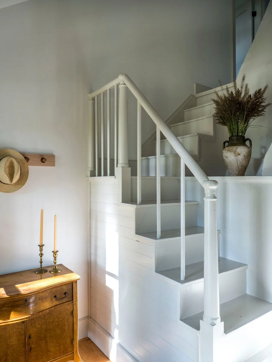 A white painted wooden staircase with horizontal wall paneling stands next to a rustic wooden cabinet topped with brass candlesticks.