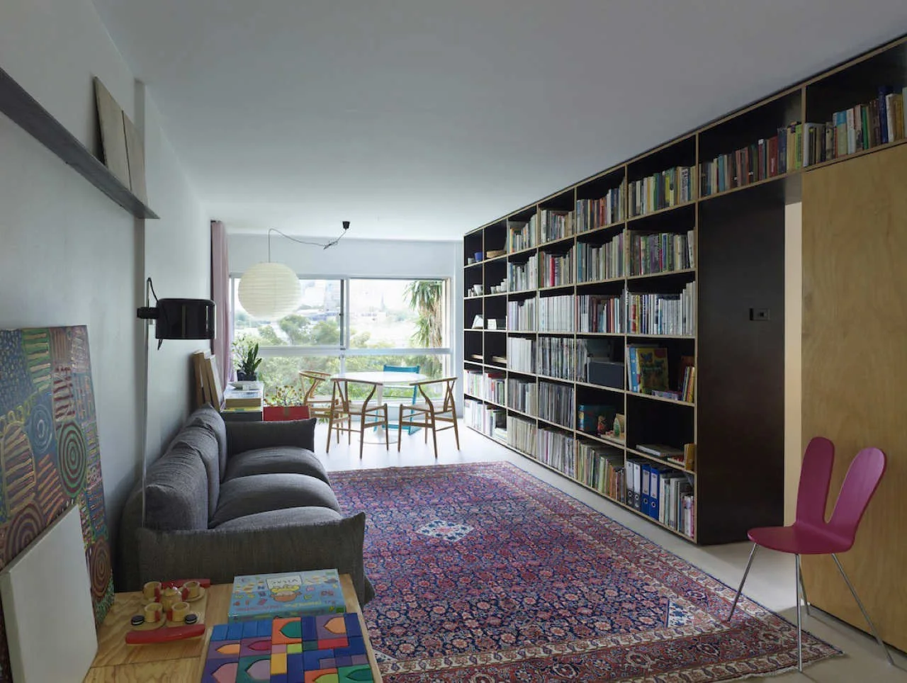 A long living room featuring a grey sofa, a large floor-to-ceiling wooden bookshelf, and a small dining set near a bright window.