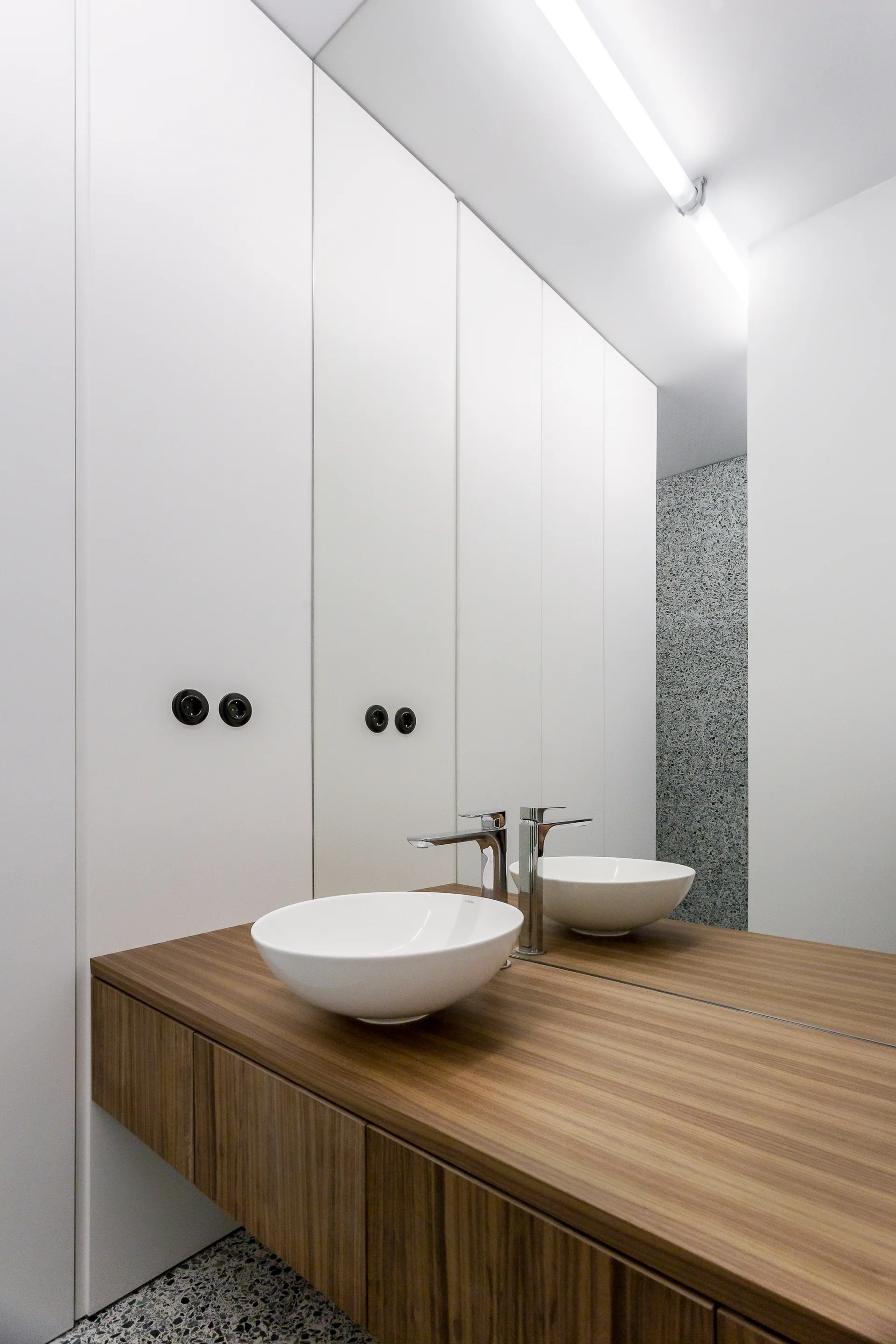 A minimalist bathroom features a floating wood vanity with a white vessel sink, a large wall mirror, and grey terrazzo flooring.