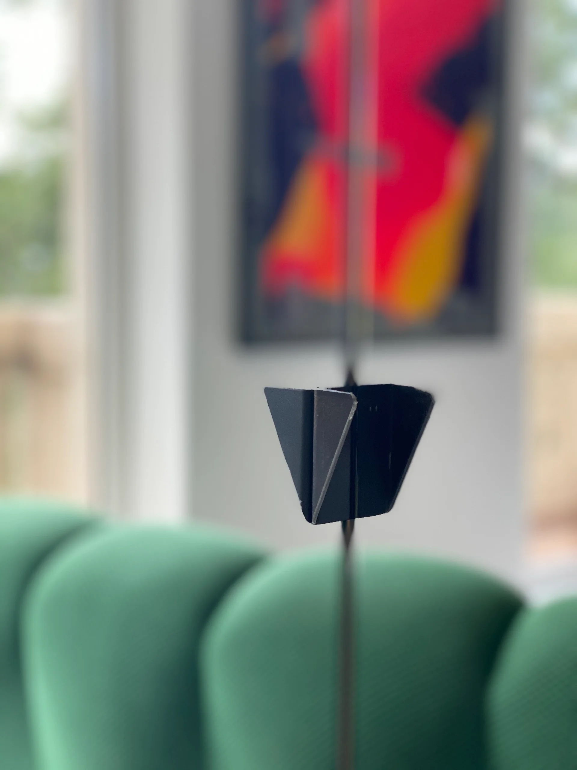 A close-up of a matte black geometric metal hardware piece with a blurred green sofa and colorful abstract artwork in the background.
