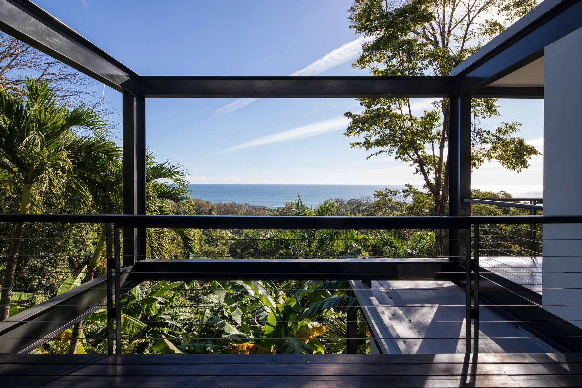 A minimalist black steel balcony with cable railing overlooks a lush green tropical forest and a blue ocean in the distance.