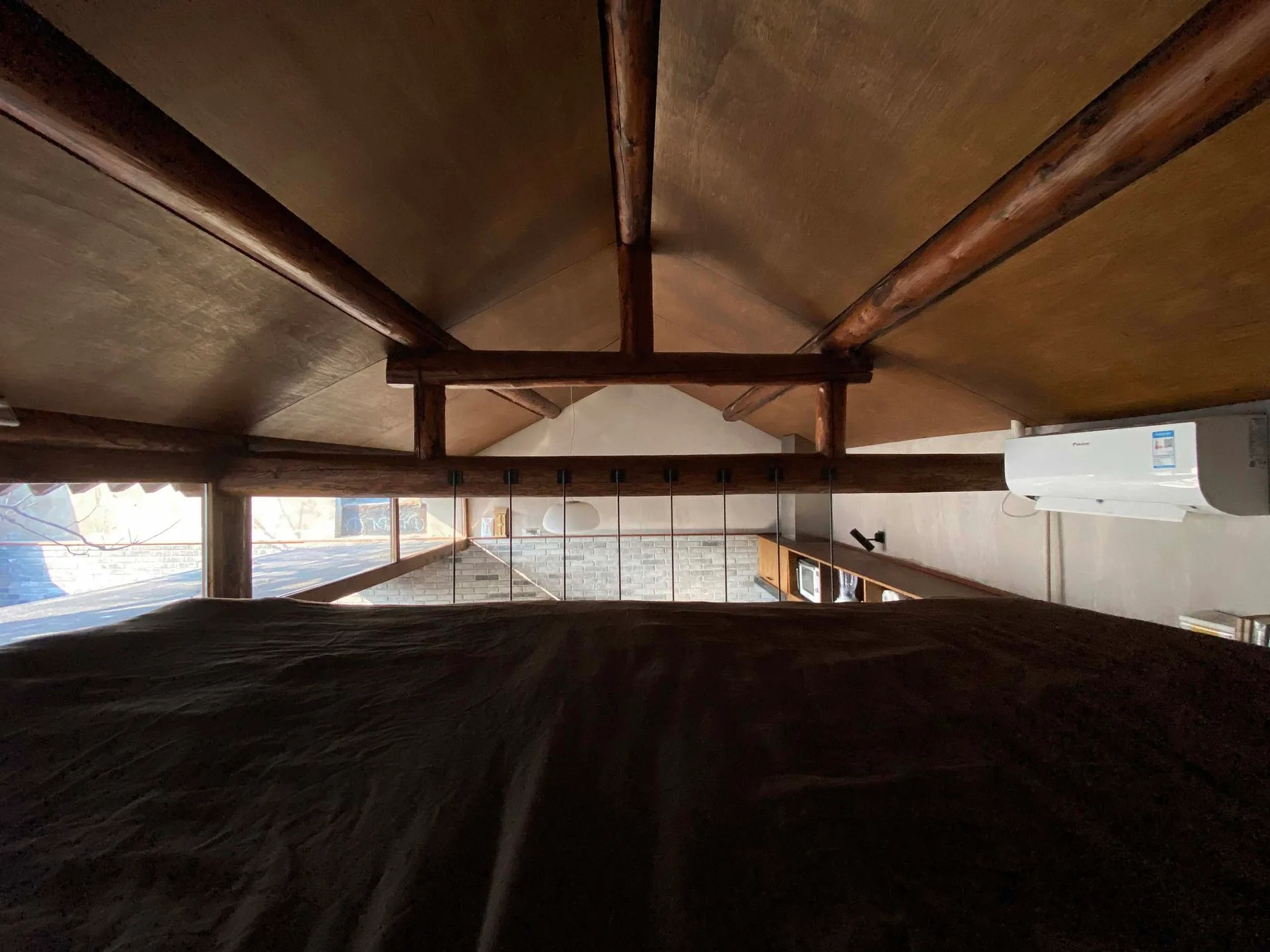 A rustic loft bedroom featuring a pitched wooden ceiling with exposed log beams overlooking a lower level with a brick wall and thin metal railing.