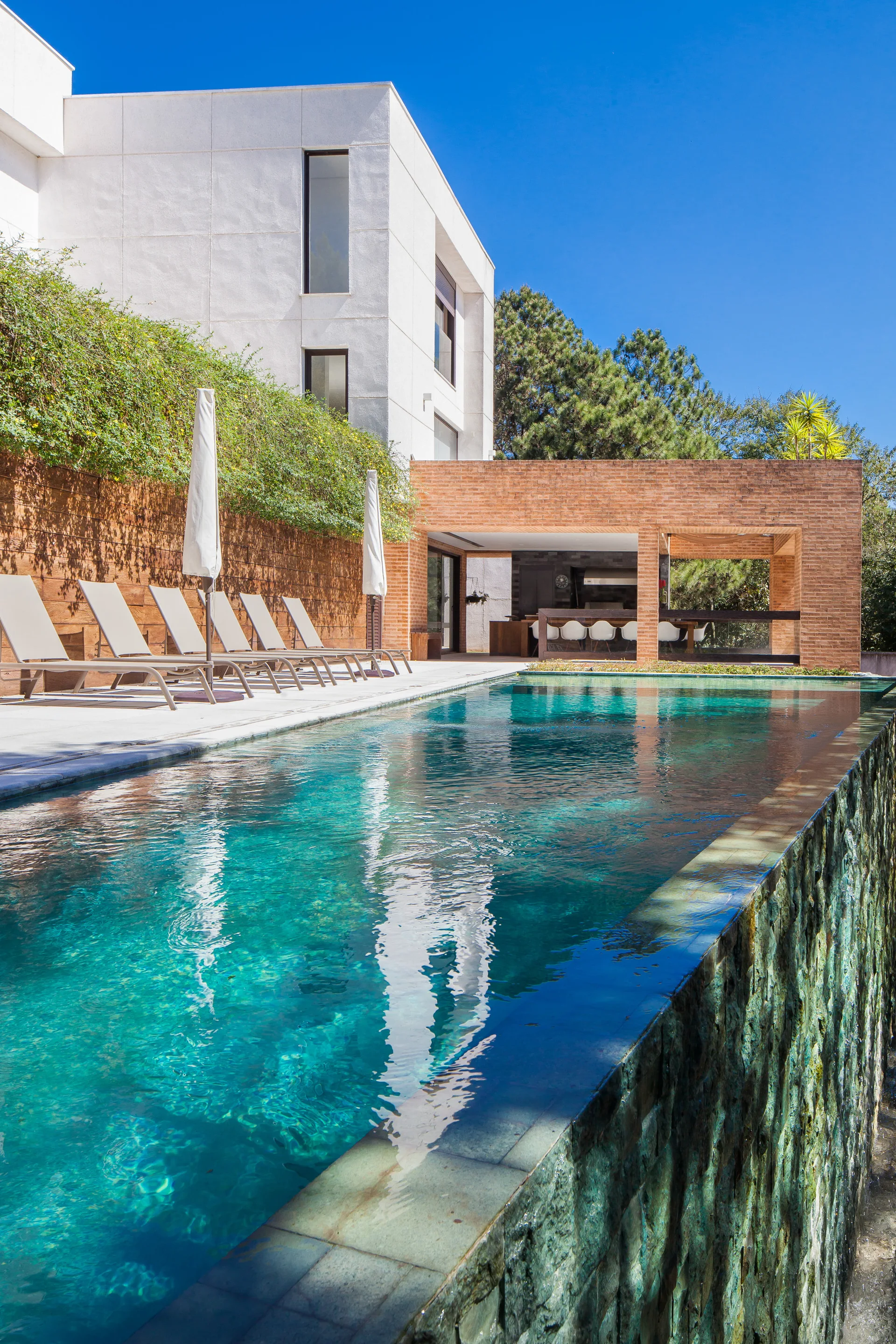 A modern infinity pool with a rustic stone outer wall features a concrete deck with lounge chairs and a white stucco house in the background.