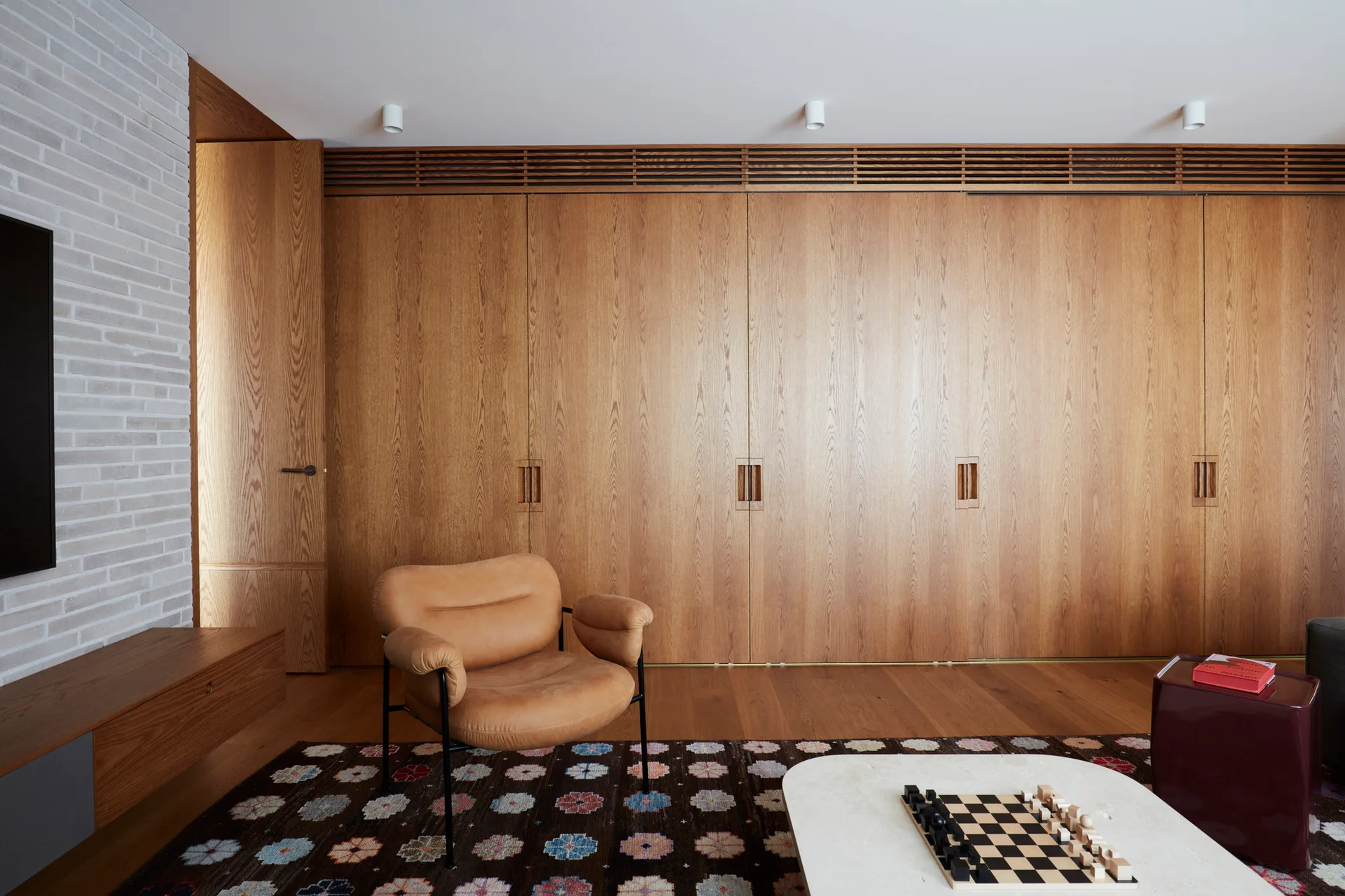 A tan leather armchair rests on a dark patterned rug in front of a floor-to-ceiling wood paneled wall.