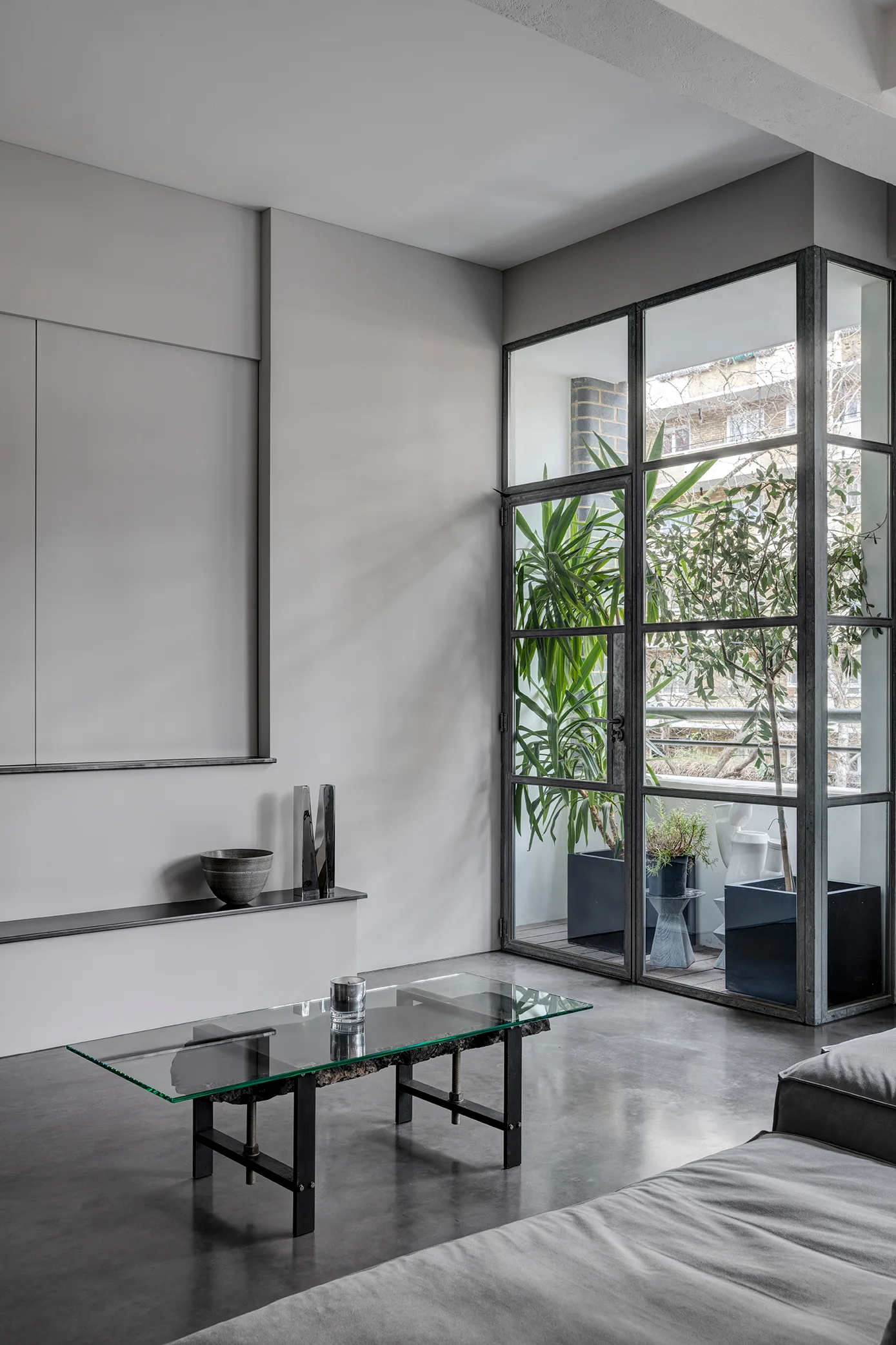 A minimalist grey living room with a glass-topped coffee table, polished concrete floors, and a large steel-framed window looking out onto a balcony.