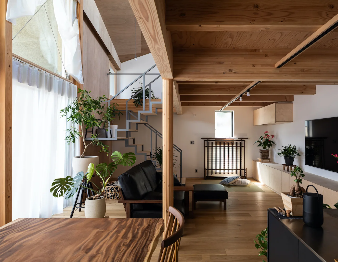 An open-concept living room features exposed wooden ceiling beams, a black leather sofa, a steel staircase, and large windows with sheer curtains.