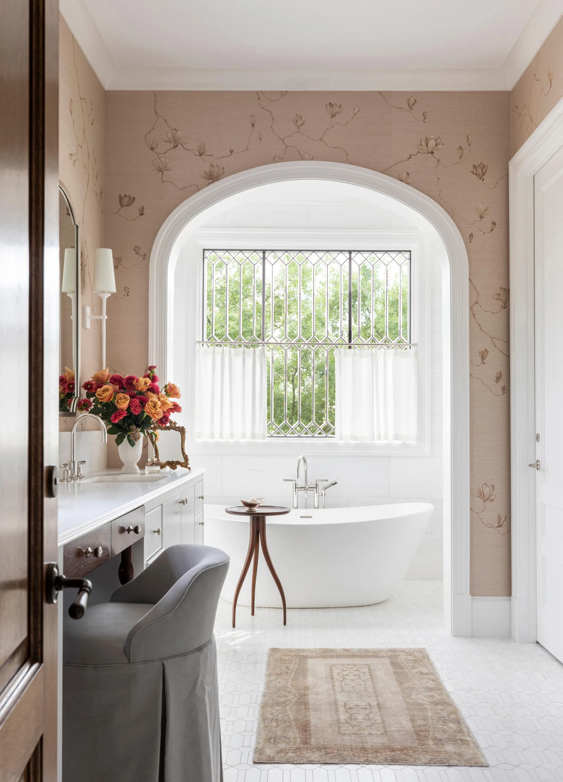 A transitional bathroom with a white freestanding tub set inside an arched alcove next to a vanity with a grey upholstered stool.