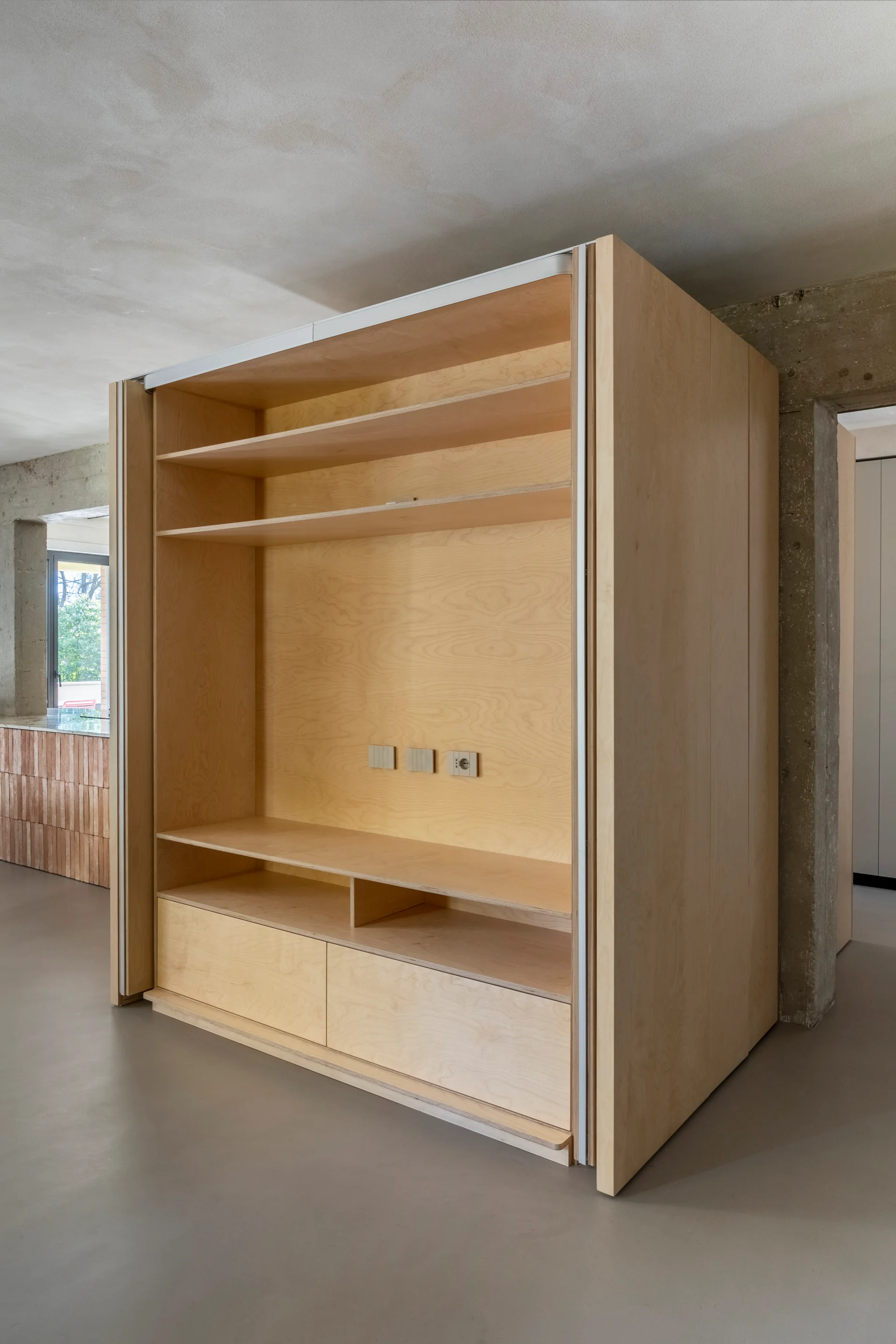 A large light plywood cabinetry unit stands on a smooth grey floor next to a raw concrete pillar under an unfinished concrete ceiling.