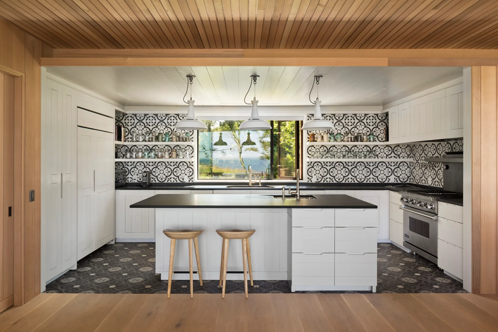 A contemporary kitchen featuring a white island, two wooden bar stools, a black and white patterned tile backsplash, and a wood plank ceiling.
