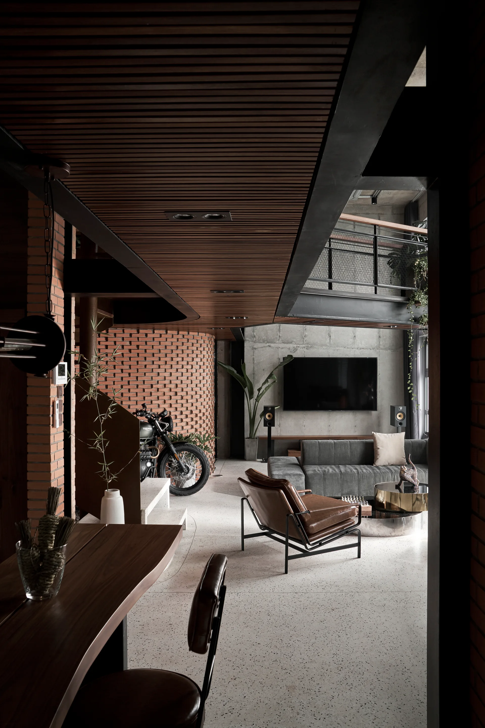 An industrial open-plan living room with a curved red brick wall, a gray sectional sofa, and a dark wood slatted ceiling over terrazzo flooring.