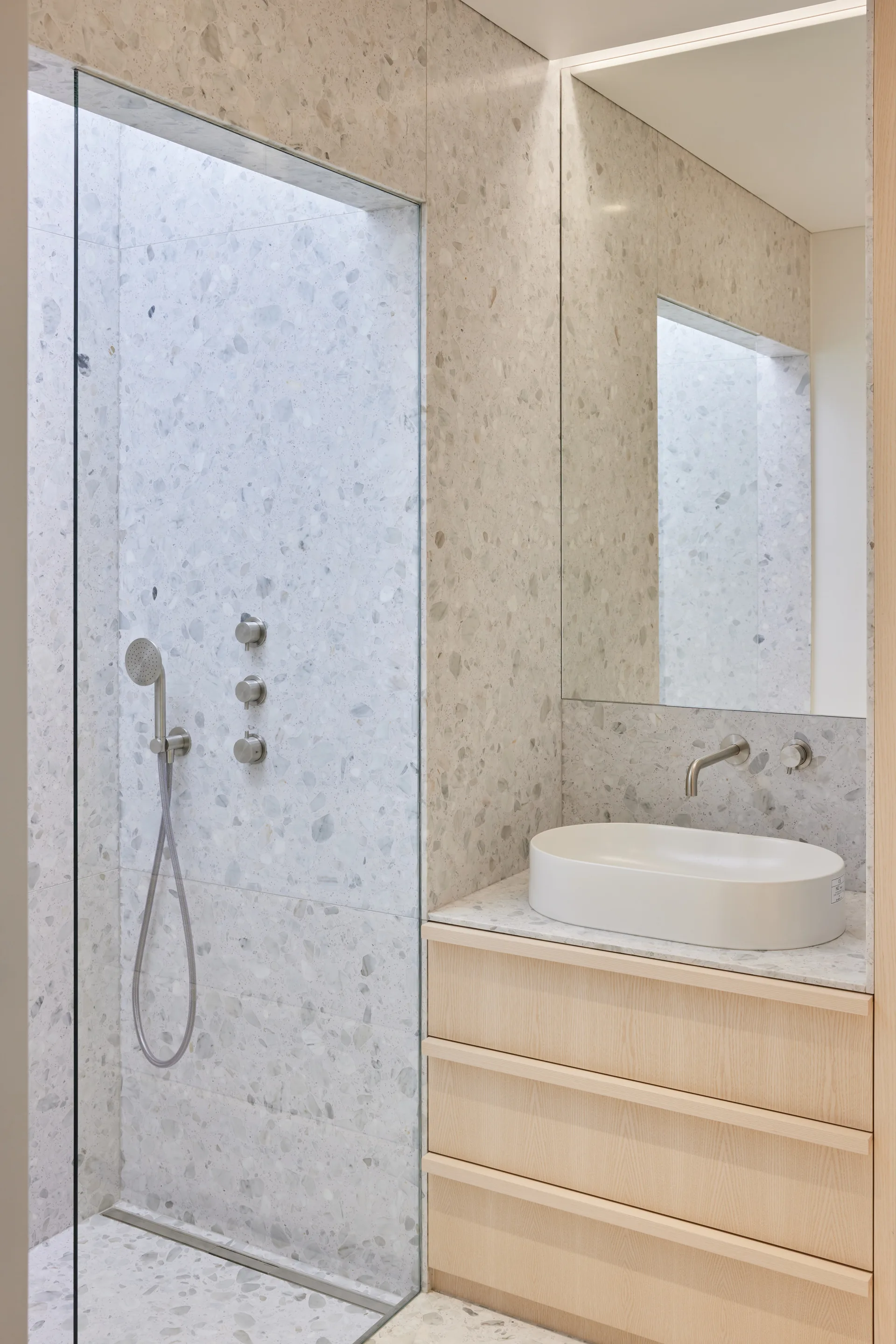 A minimalist bathroom features a light wood vanity with an oval vessel sink, a wall-mounted faucet, and a walk-in shower with grey terrazzo tile walls.