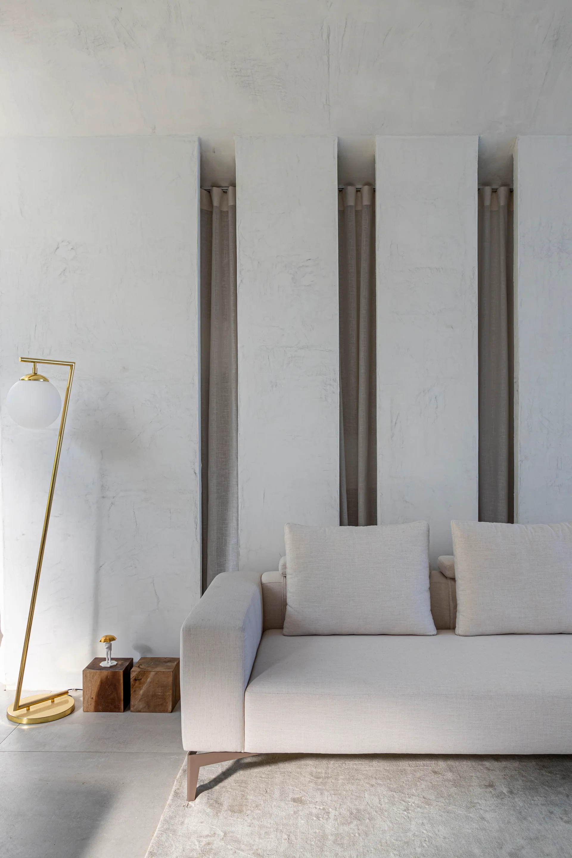 A cream fabric sofa sits in front of a textured plaster wall featuring deep vertical slots with recessed neutral curtains beside a gold globe floor lamp.
