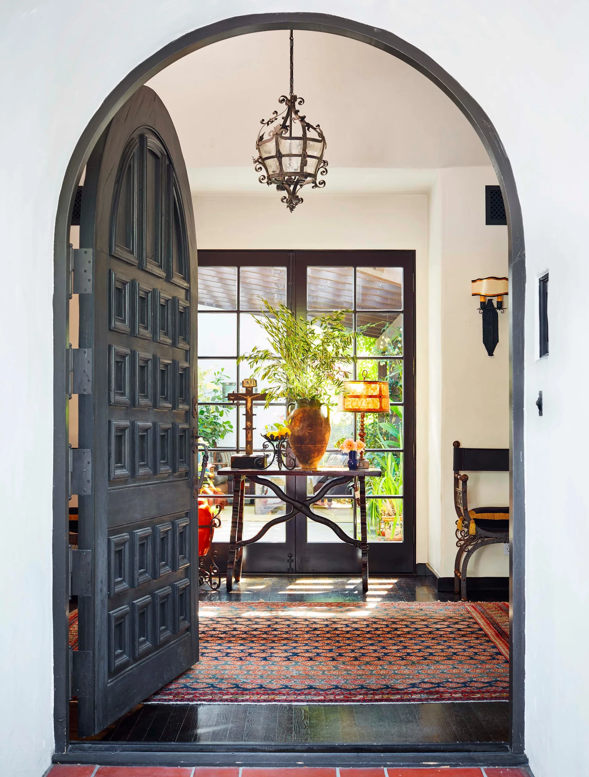 A Spanish Colonial entryway features a large black arched door standing open, revealing a patterned rug, a console table, and glass French doors.