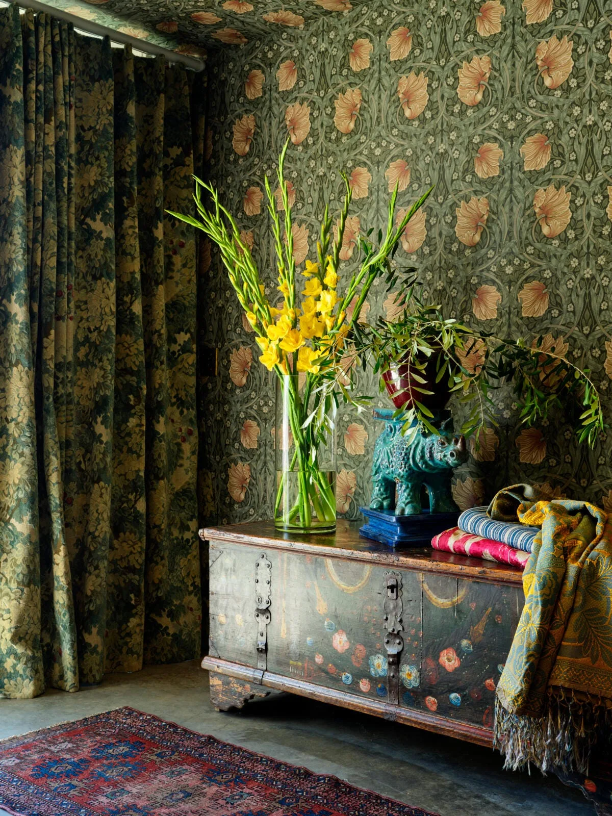 An antique painted wooden chest sits in a corner covered in green botanical wallpaper, holding tall yellow gladiolus flowers and a small ceramic sculpture.