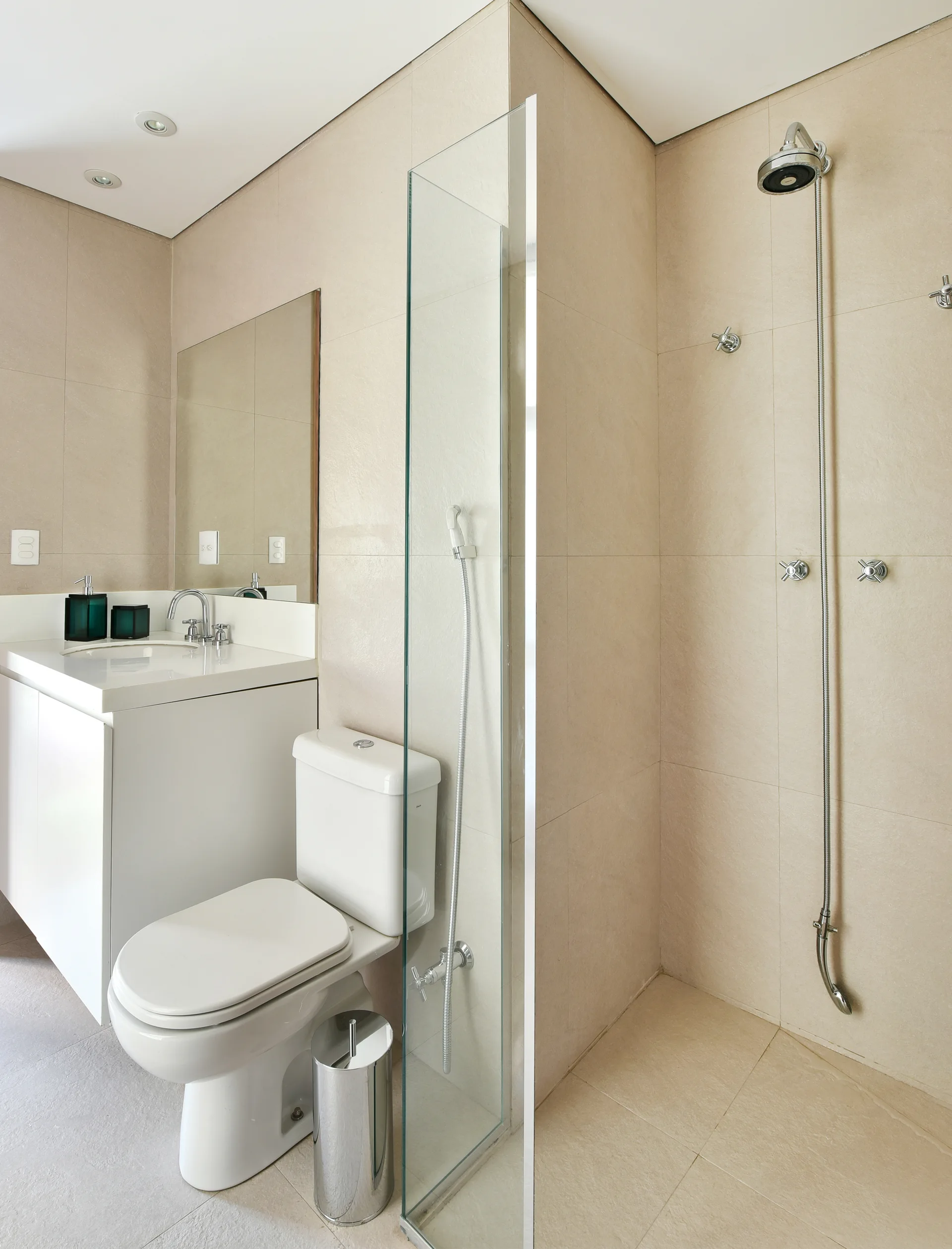 A modern bathroom features beige tiled walls and floors, a white vanity with a mirror, a toilet, and a walk-in shower with a clear glass panel.