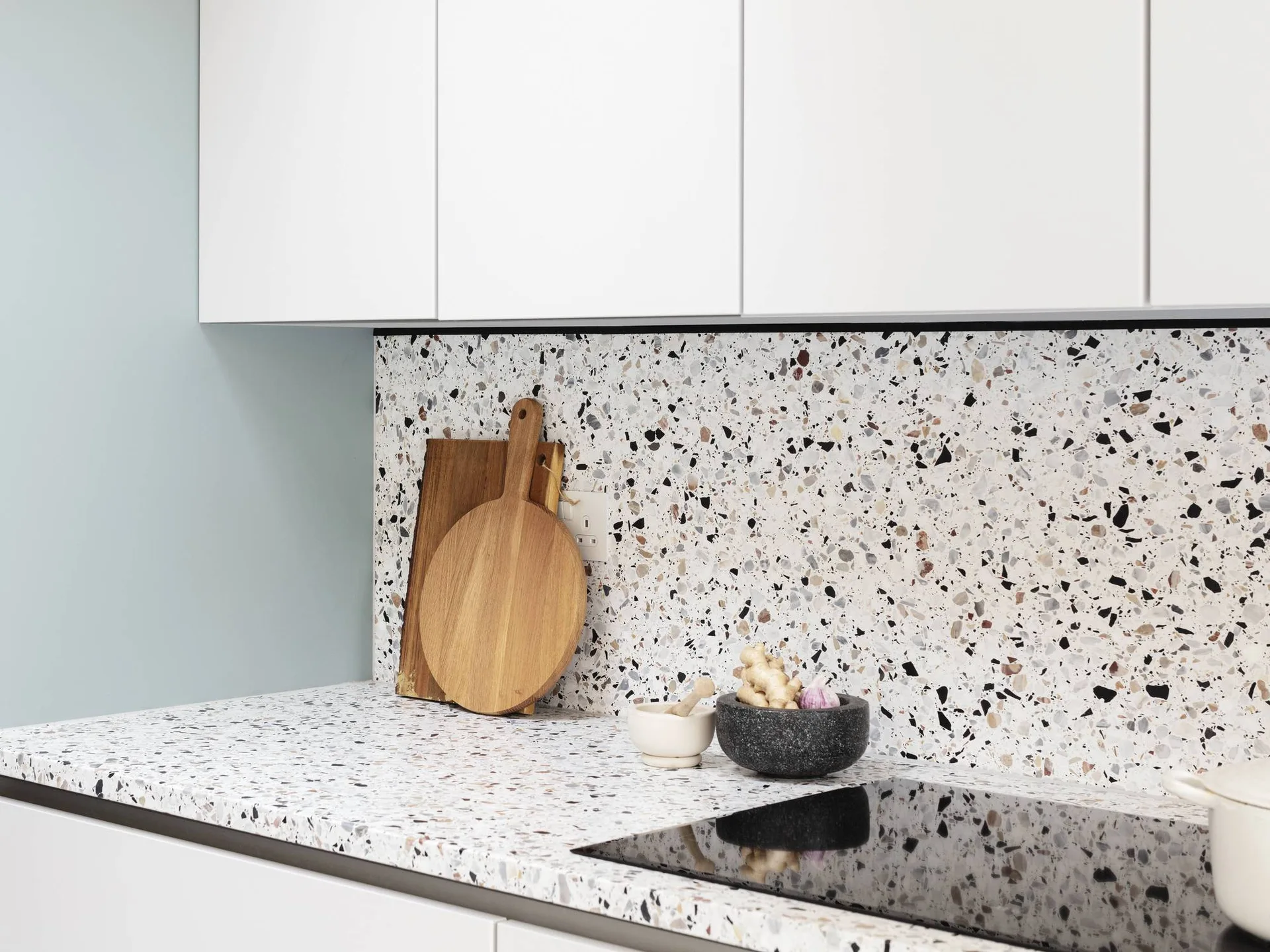 A modern kitchen featuring matte white flat-front cabinets above a matching terrazzo stone countertop and backsplash with small speckled patterns.