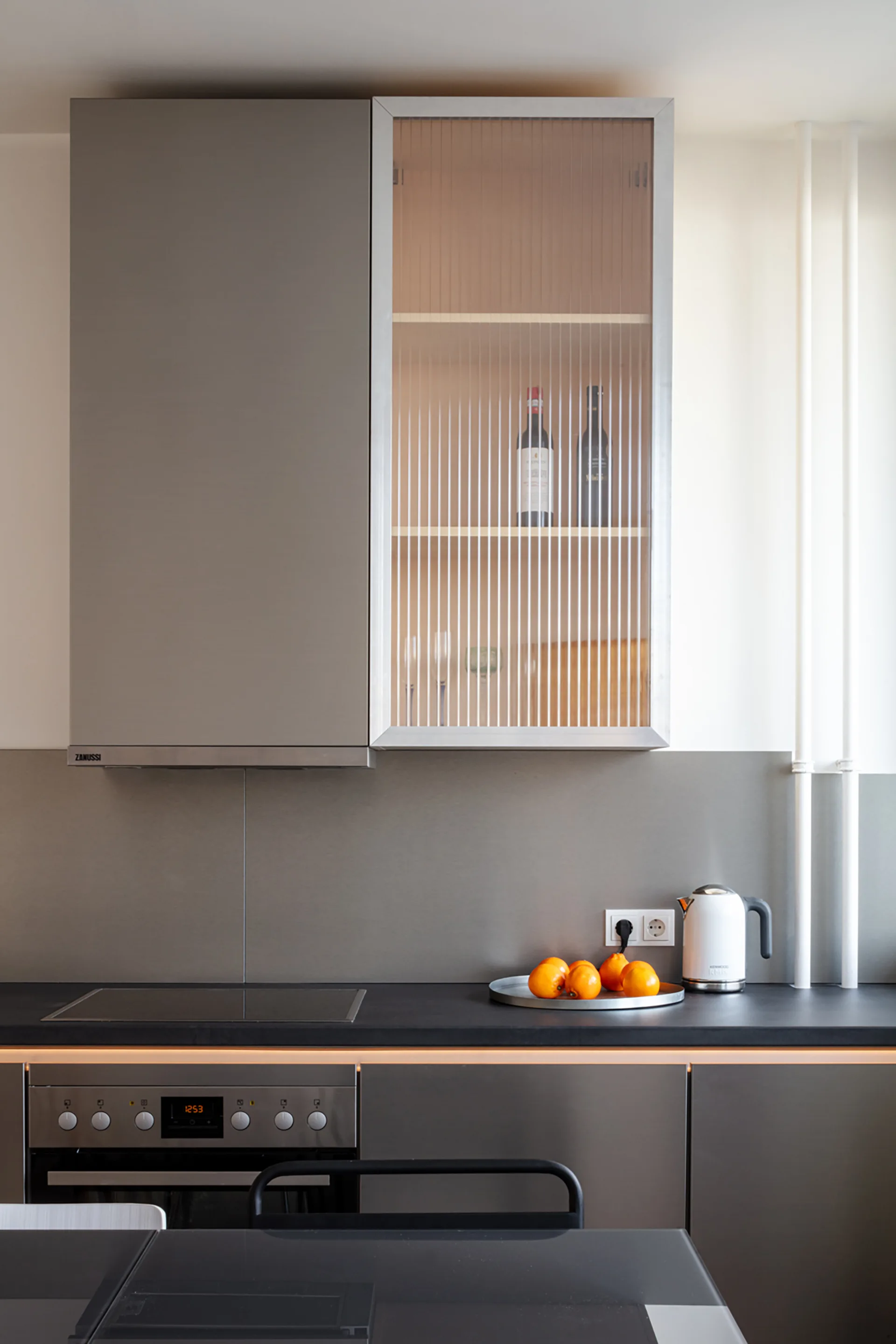 A modern kitchen featuring matte grey cabinets, a reeded glass upper cabinet door, under-cabinet lighting, and a black countertop with oranges.