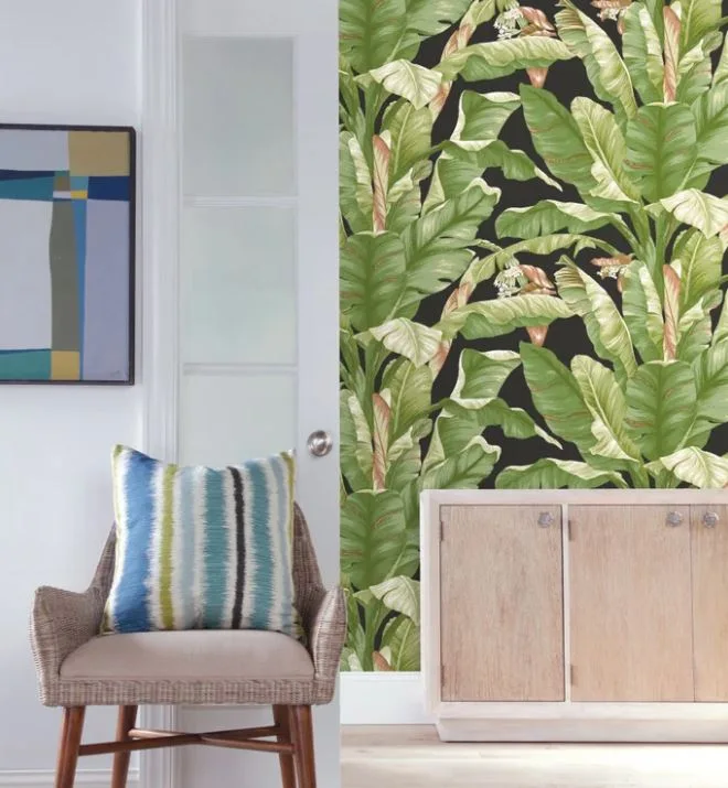 An entryway corner features a wicker armchair with a striped pillow next to a white door, with green tropical leaf wallpaper and a light wood cabinet.