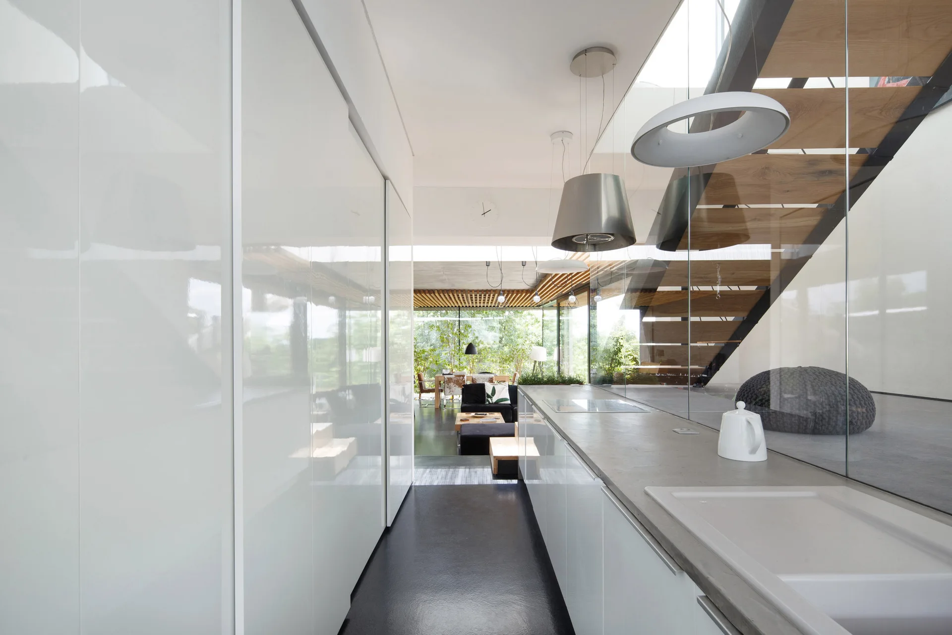 A modern galley kitchen features high-gloss white cabinets, a grey countertop, and a glass partition separating it from a wood-tread staircase.