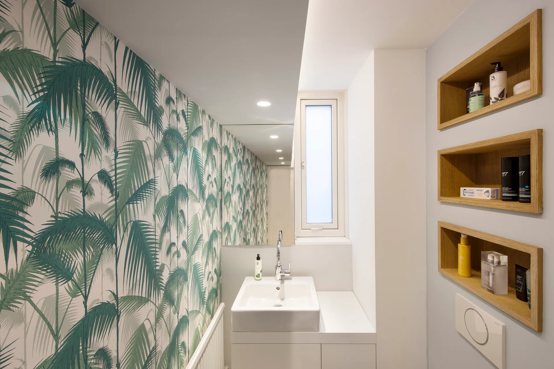 A compact modern bathroom features green palm-leaf wallpaper, a white vanity sink, and three recessed wooden shelves filled with toiletries.
