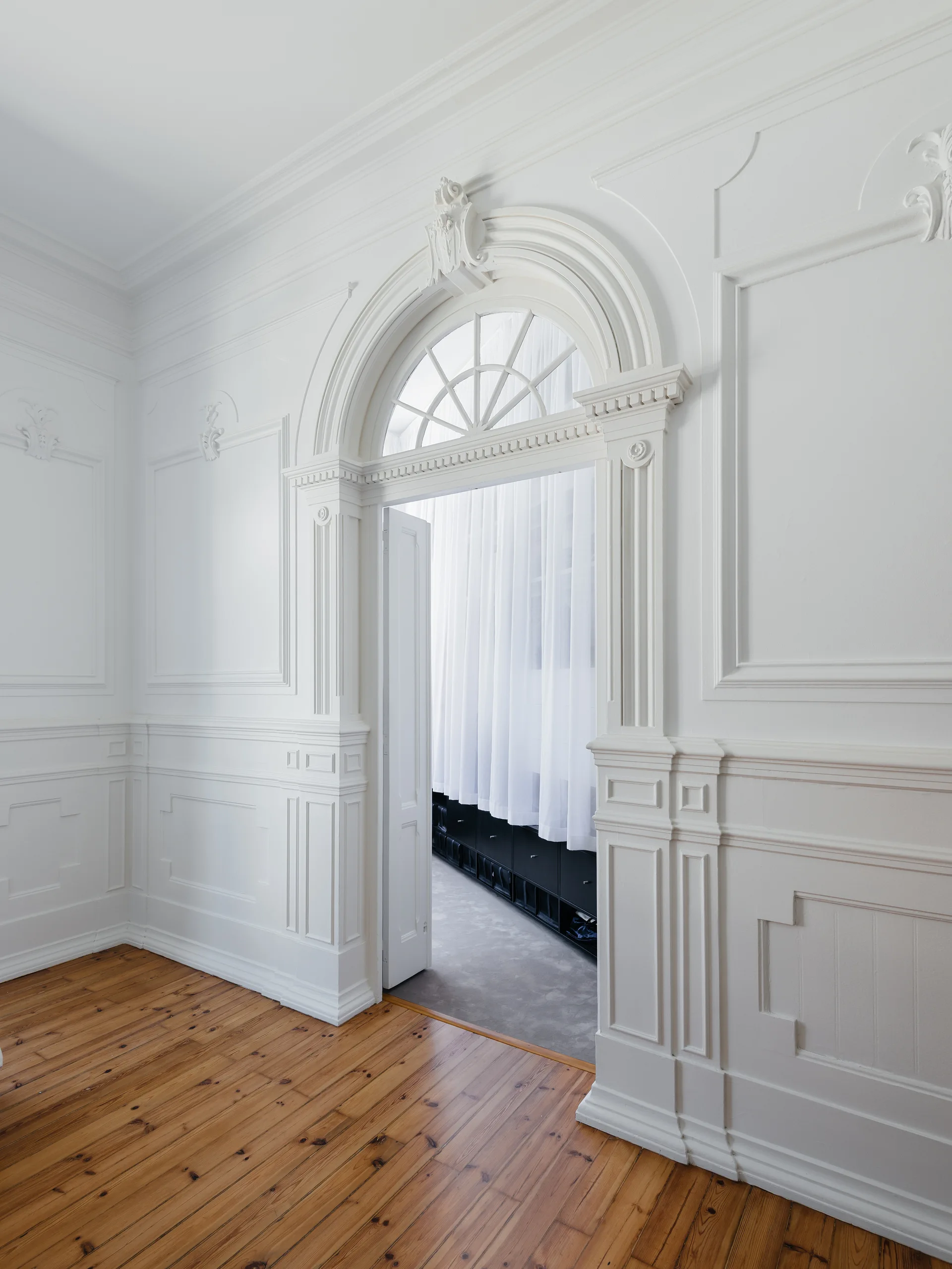 An ornate white archway with a glass transom window opens into an adjacent room, framed by detailed wall paneling and warm wood floors.