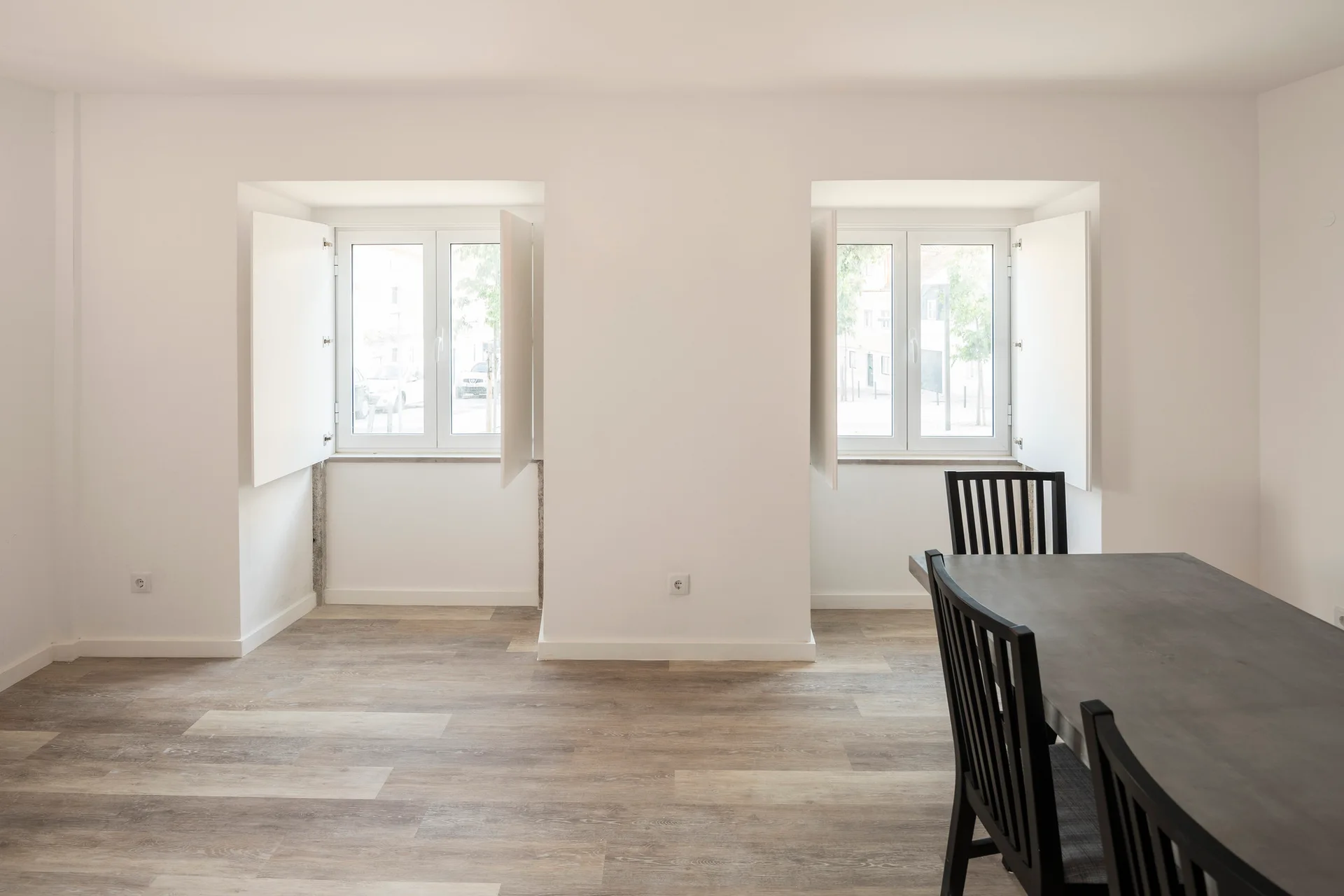 A minimalist dining room features a dark wood table with black slat-back chairs alongside two symmetrical recessed windows with white folding shutters.