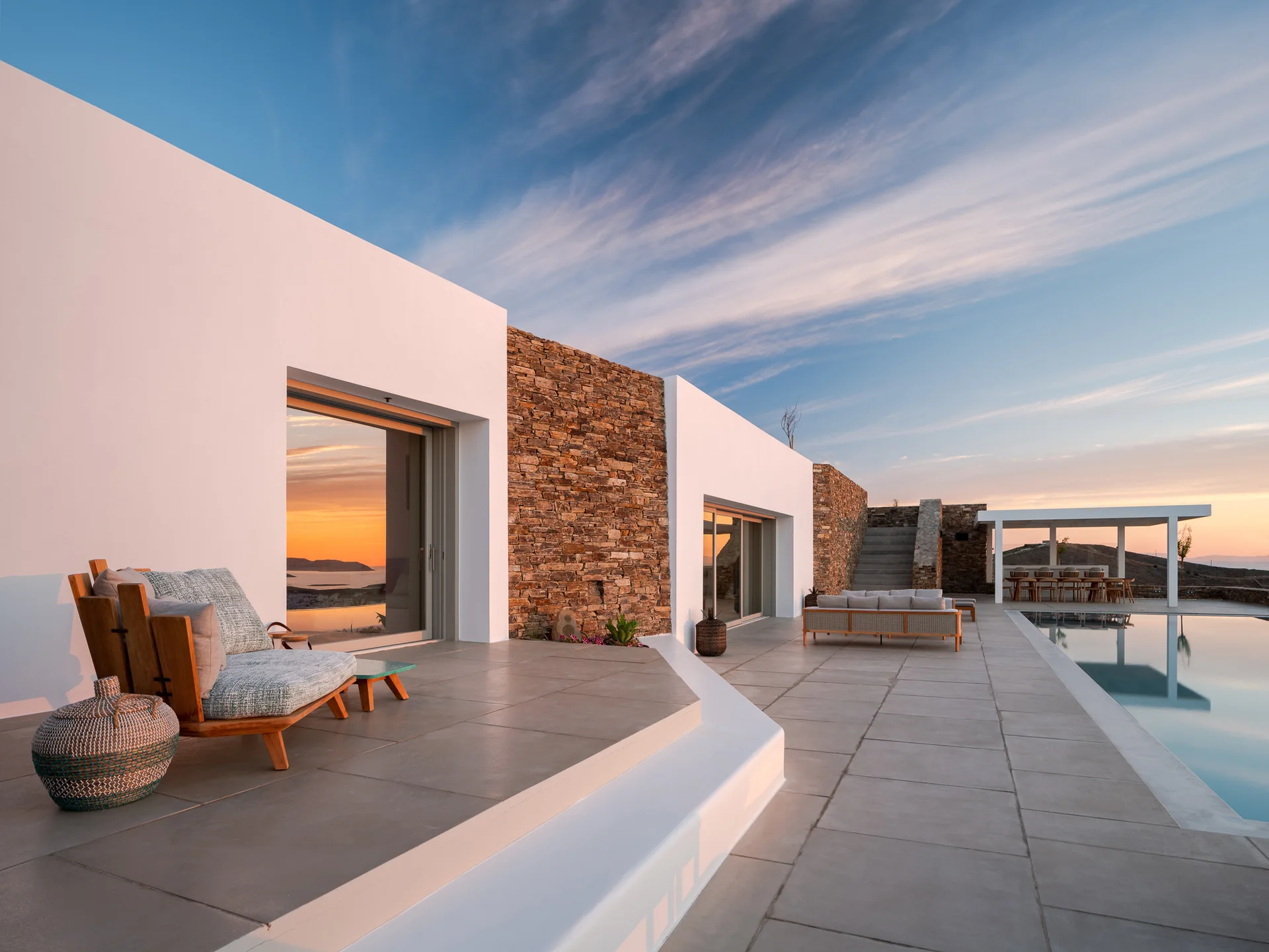 A modern outdoor patio features a stone and white stucco exterior, an infinity pool, wood lounge furniture, and views of a sunset.