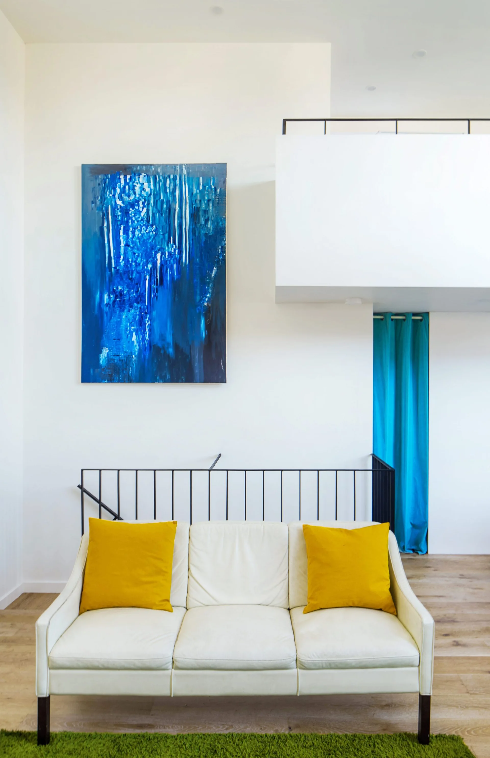A white leather-like sofa with two yellow pillows sits in front of a black metal railing and a large blue abstract painting on a white wall.