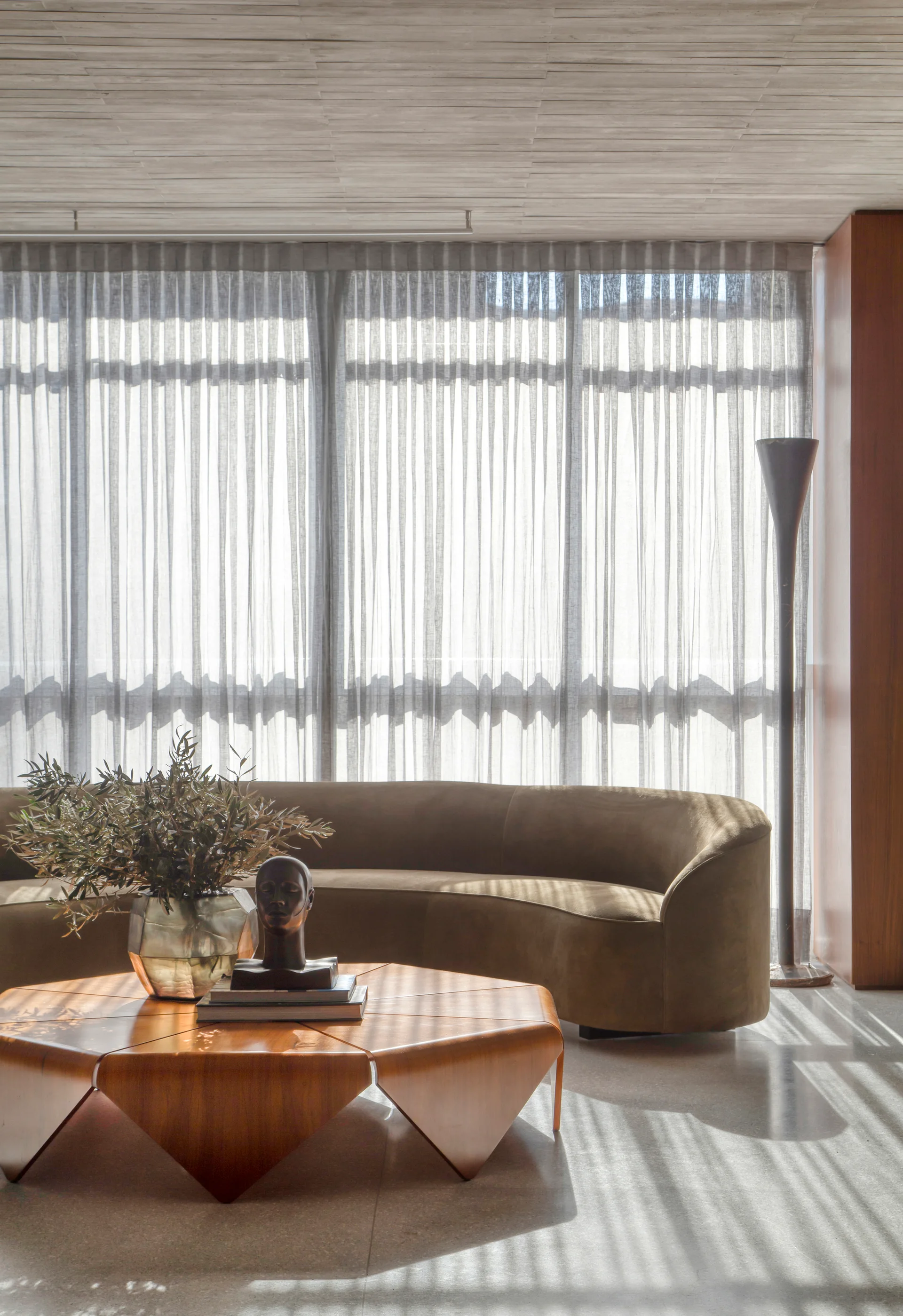 A curved brown sofa and a geometric wooden coffee table are set against floor-to-ceiling sheer curtains filtering bright natural light.