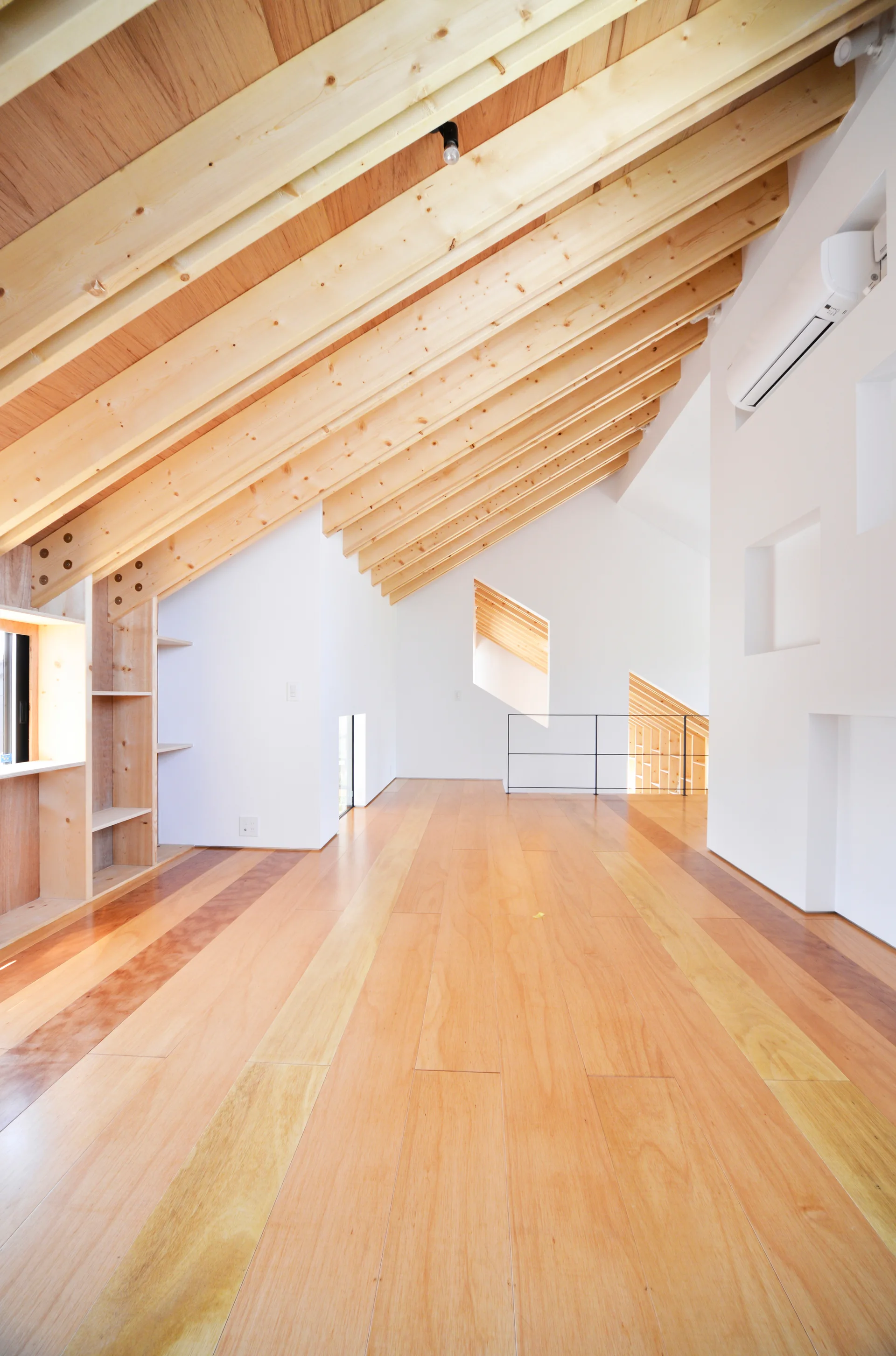 A spacious loft landing features a steeply sloped ceiling with exposed timber rafters, white walls, and multi-toned wood plank flooring.