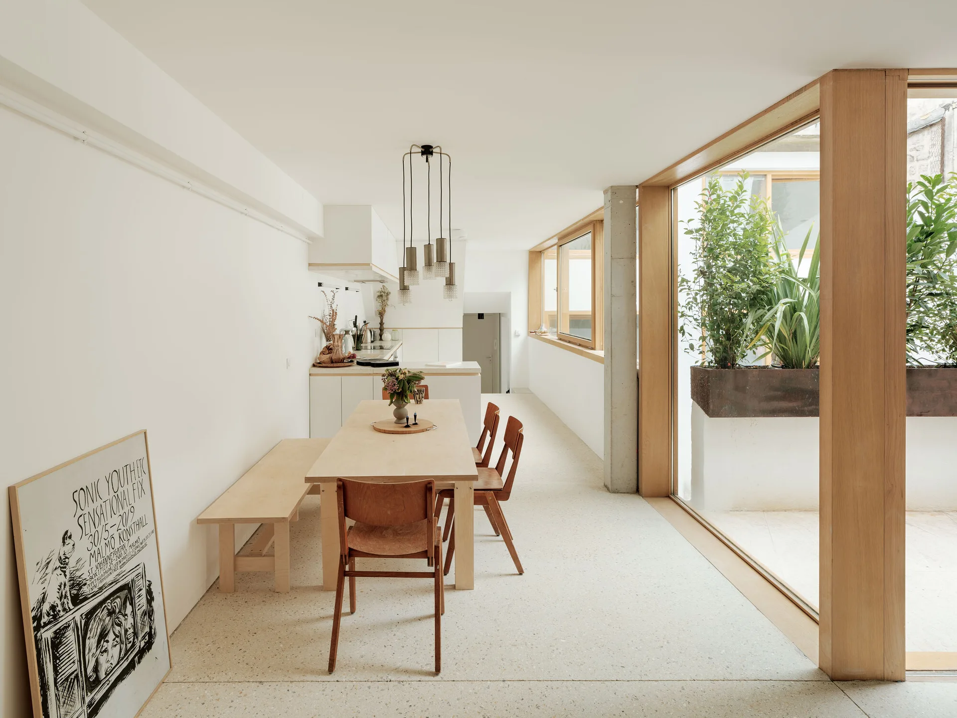 A minimalist dining area features a light wood table, bench, and chairs set on speckled terrazzo flooring next to large glass windows.