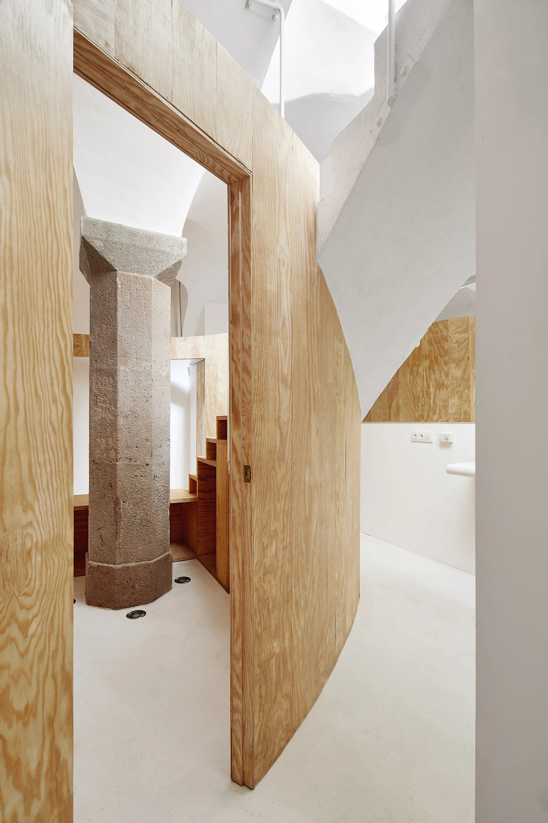 A minimalist interior featuring curved plywood partition walls, an octagonal stone column, white vaulted ceilings, and smooth white floors.