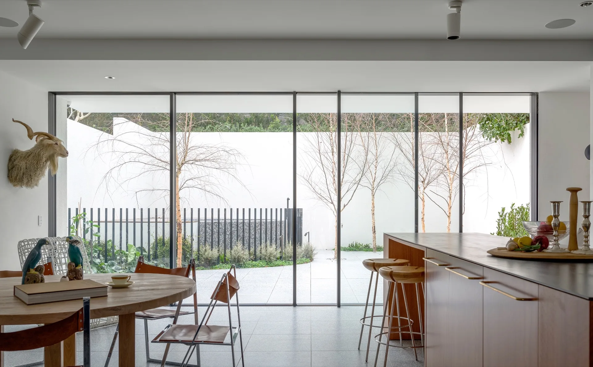 A modern kitchen and dining area featuring a wooden island, circular table, and large sliding glass doors opening to a minimalist courtyard.